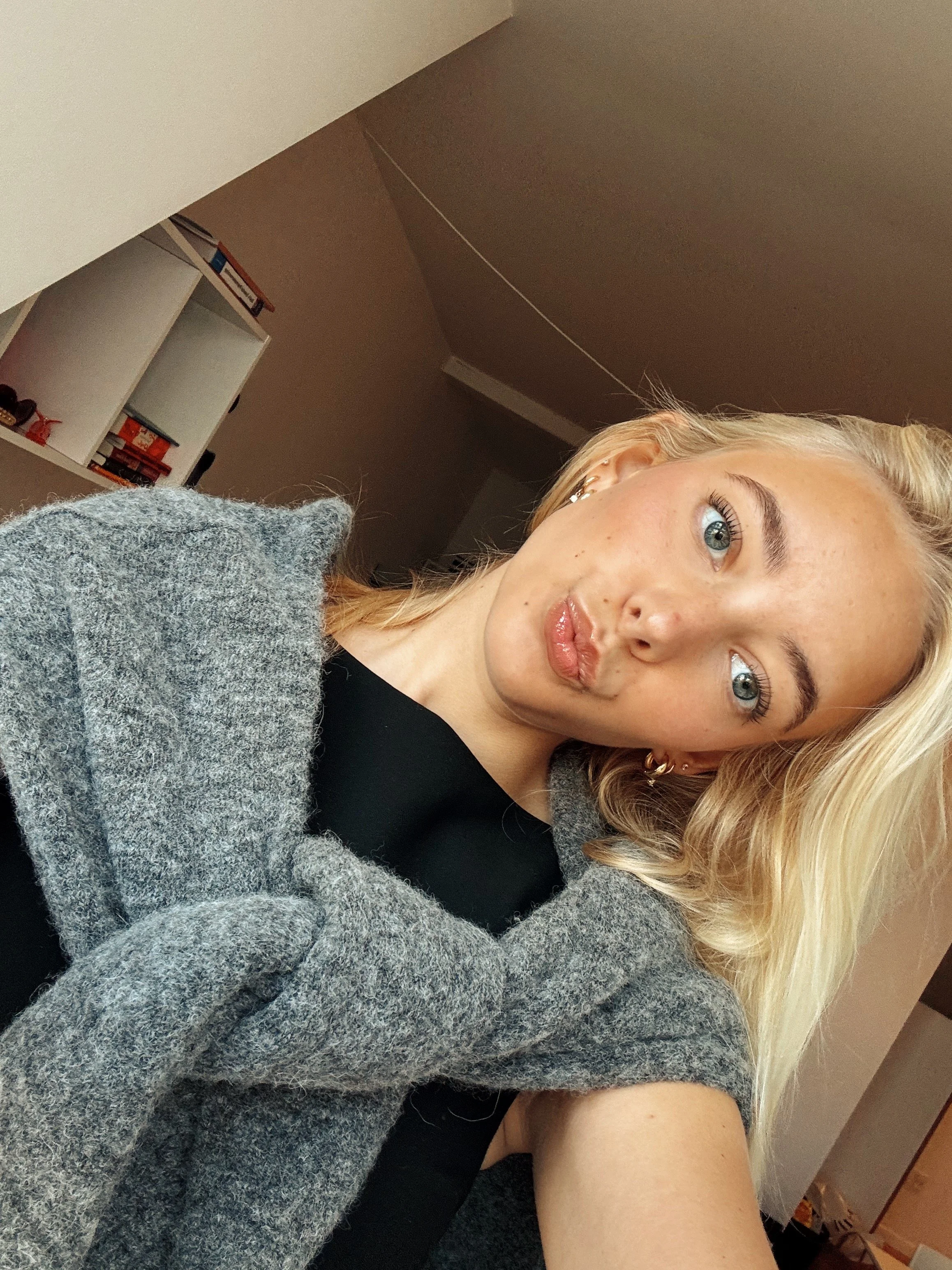 Close-up of a young woman with blond hair and blue eyes taking a selfie indoors, wearing a gray cardigan over a black top.