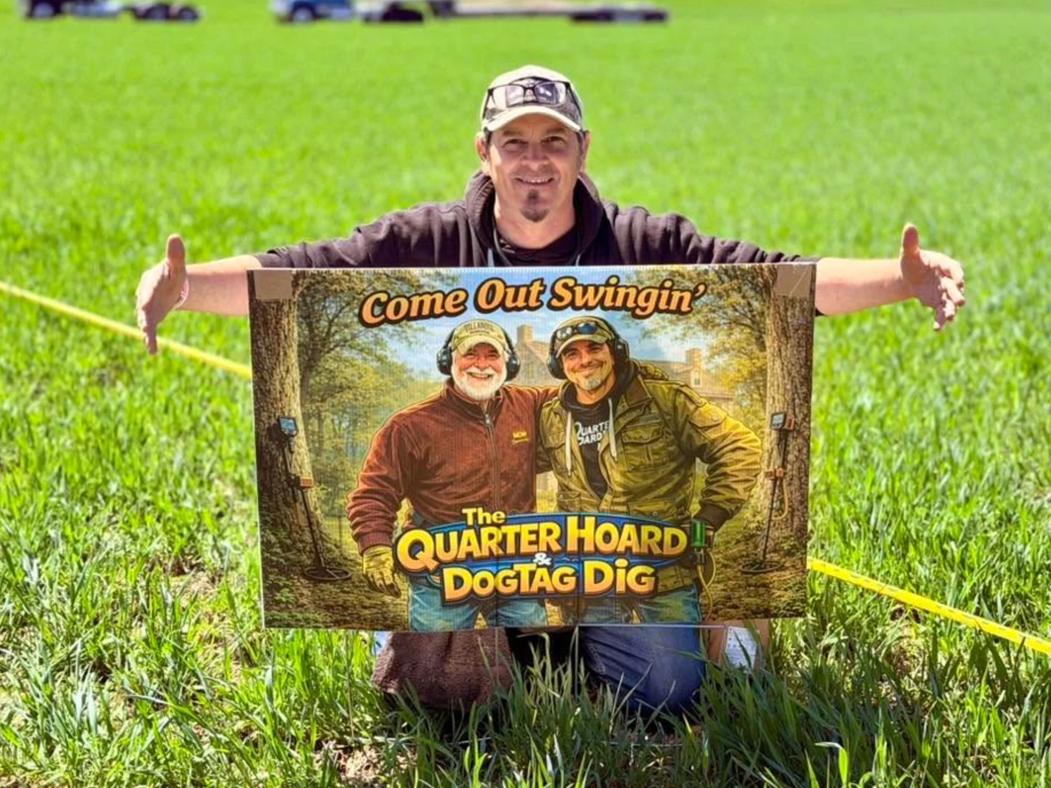 Jason from Quarter Hoarder kneeling behind a sign that says 'The Quarter Hoard & Dog Tag Dig', a hunt put on at the Come Out Swinging metal detecting event in 2026
