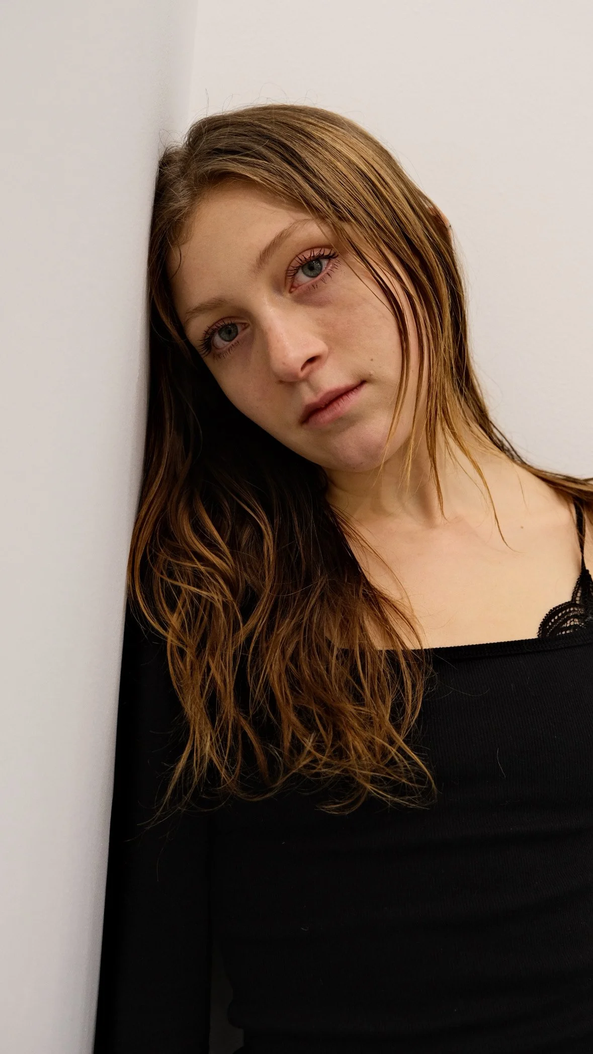A young woman with long, wavy auburn hair and blue eyes leaning against a white wall, wearing a black top.