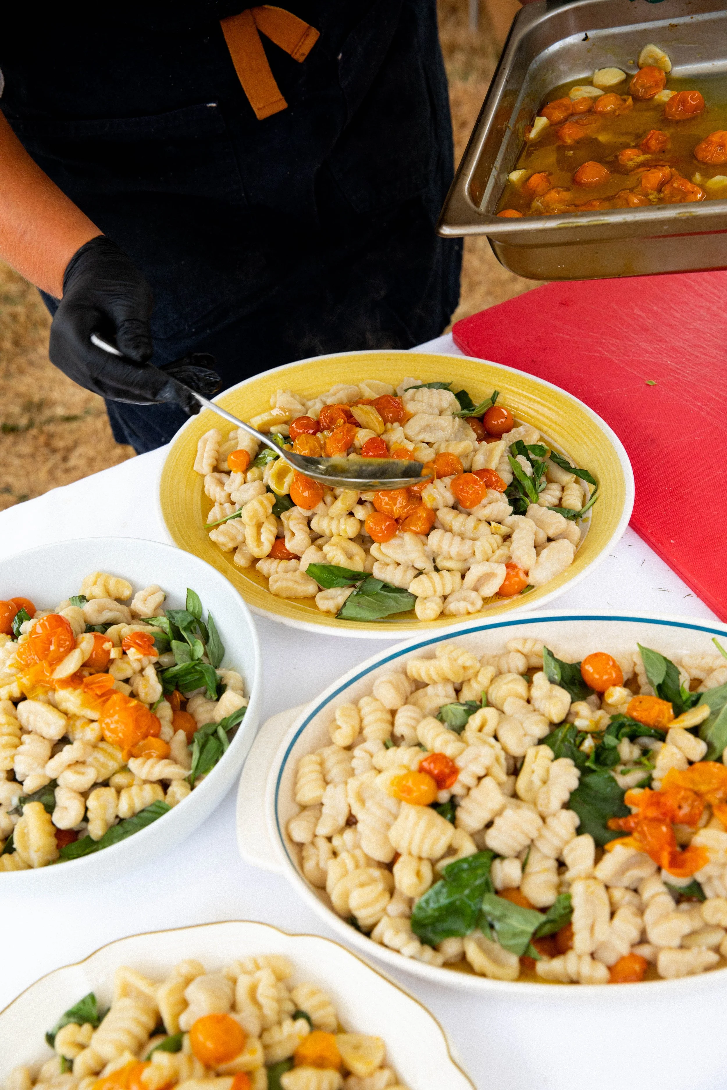 A person in black gloves is serving pasta salad with cherry tomatoes, basil, and green peppers from a large tray onto a yellow plate. Multiple plates of the same pasta salad are on the table.