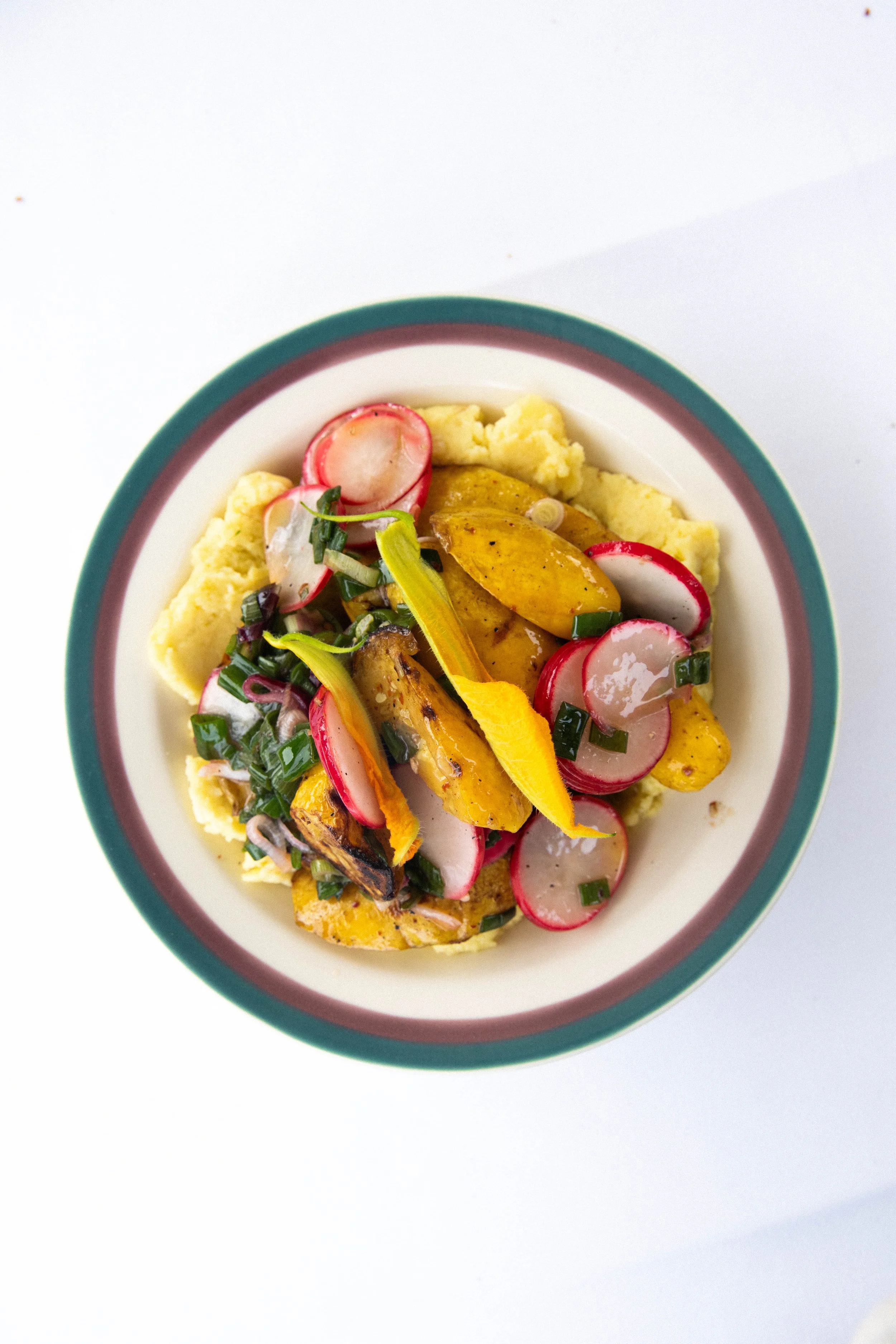 A plate of roasted vegetables and mashed potatoes, garnished with sliced radishes, chopped green onions, and yellow bell pepper strips, on a white background.
