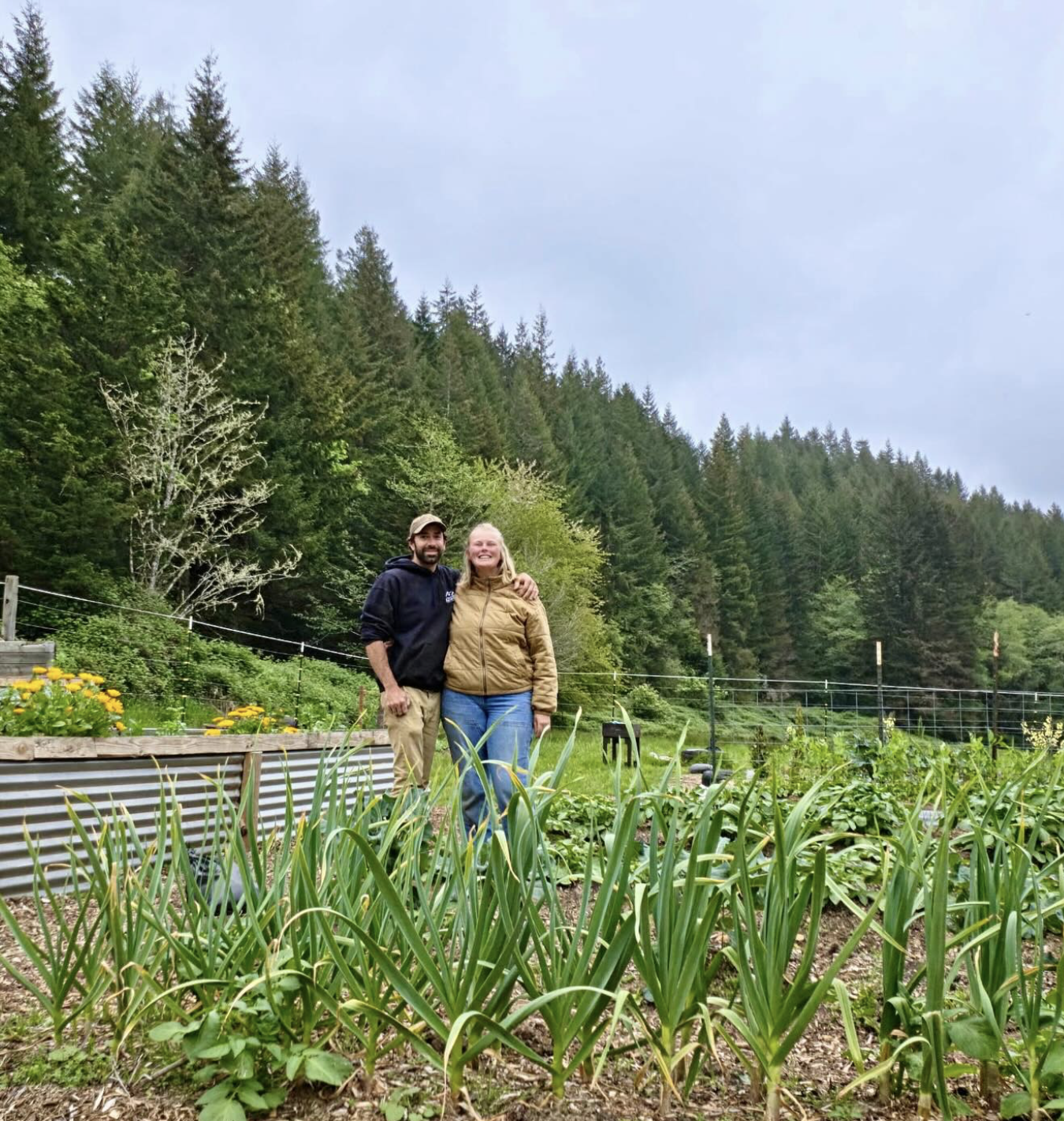 A man and woman smiling and standing together in a garden with lush green plants, yellow flowers, and a backdrop of tall trees and a cloudy sky.