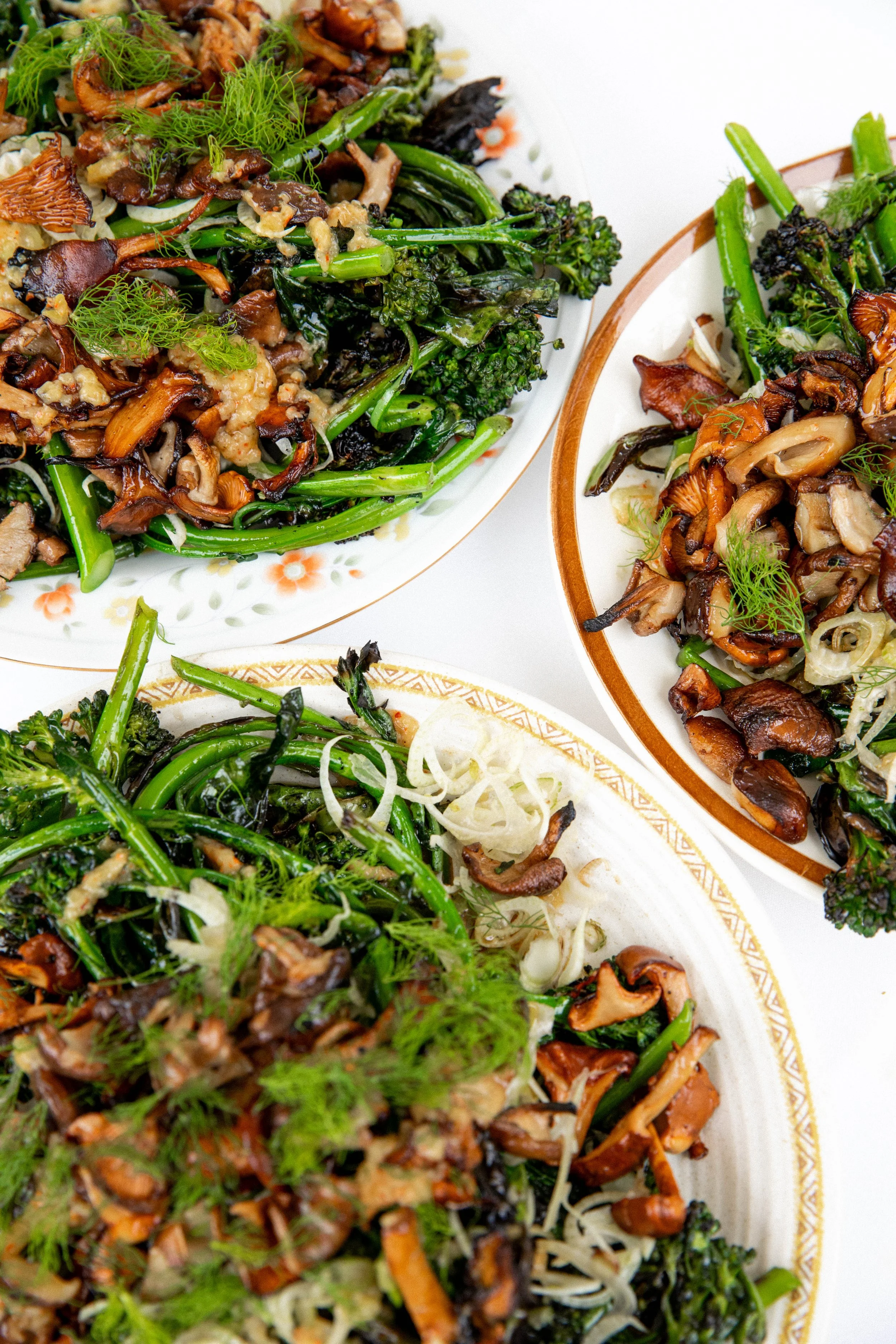 Close-up of various sautéed vegetables on decorative plates, including broccoli, mushrooms, and green beans, garnished with fresh herbs.