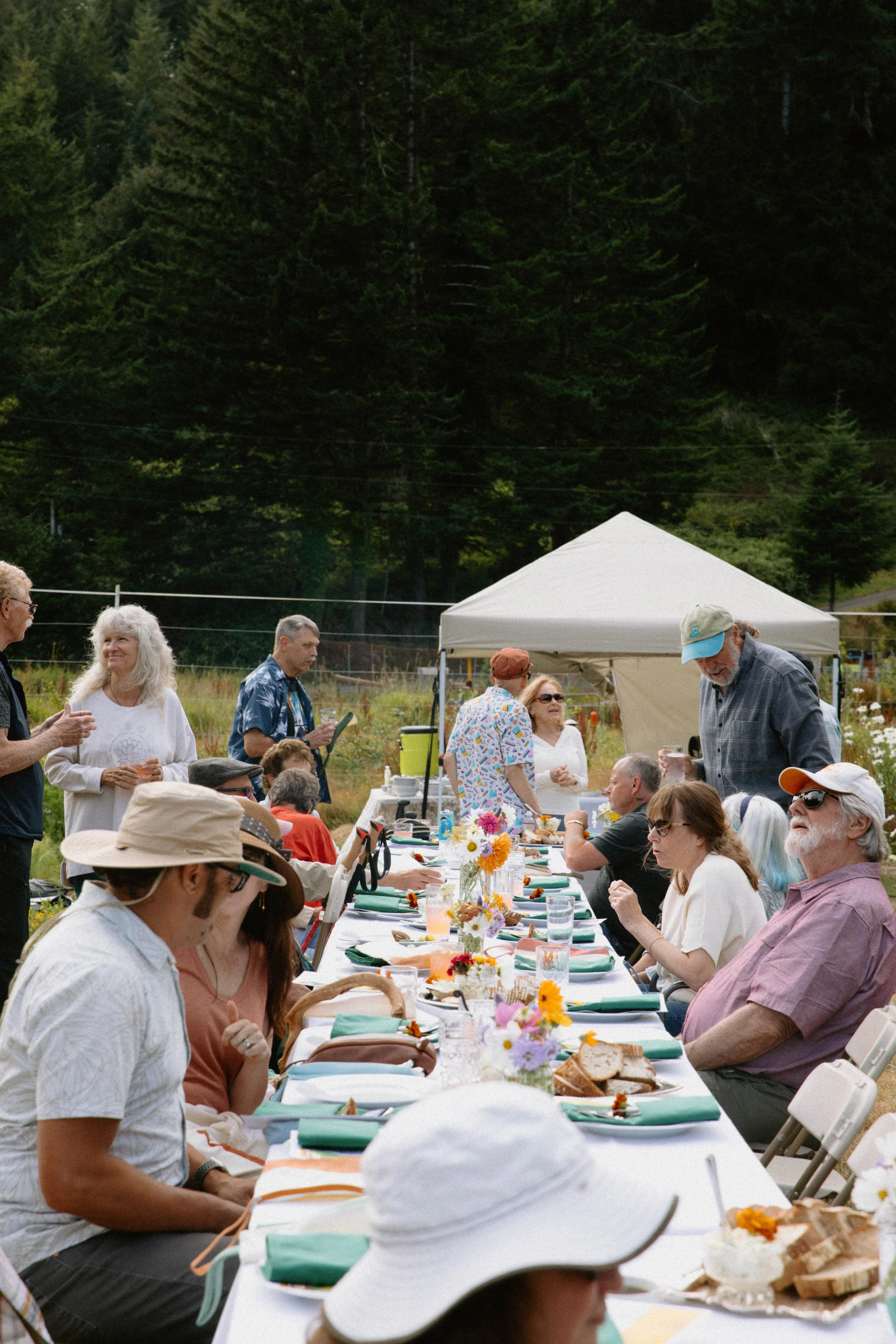 People gather outdoors around a long decorated table, with some standing and others sitting, in a lush green setting with trees in the background, under a white tent.