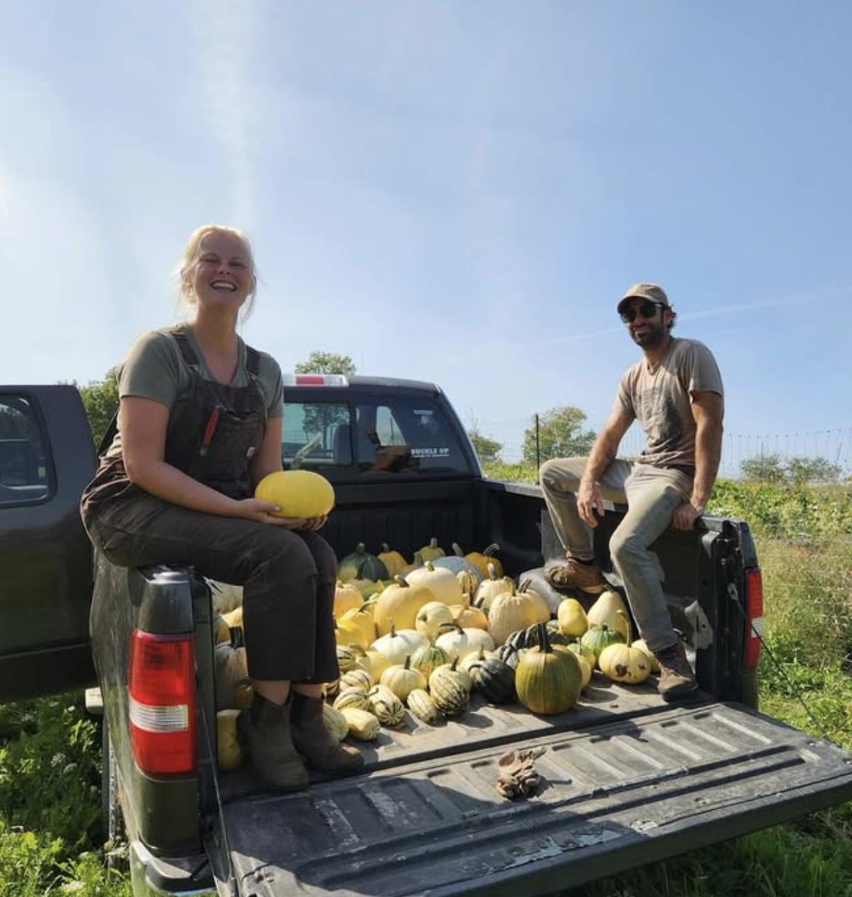 A woman and a man sitting on the back of a pickup truck filled with yellow and green gourds, in an outdoor farm setting under a clear blue sky.