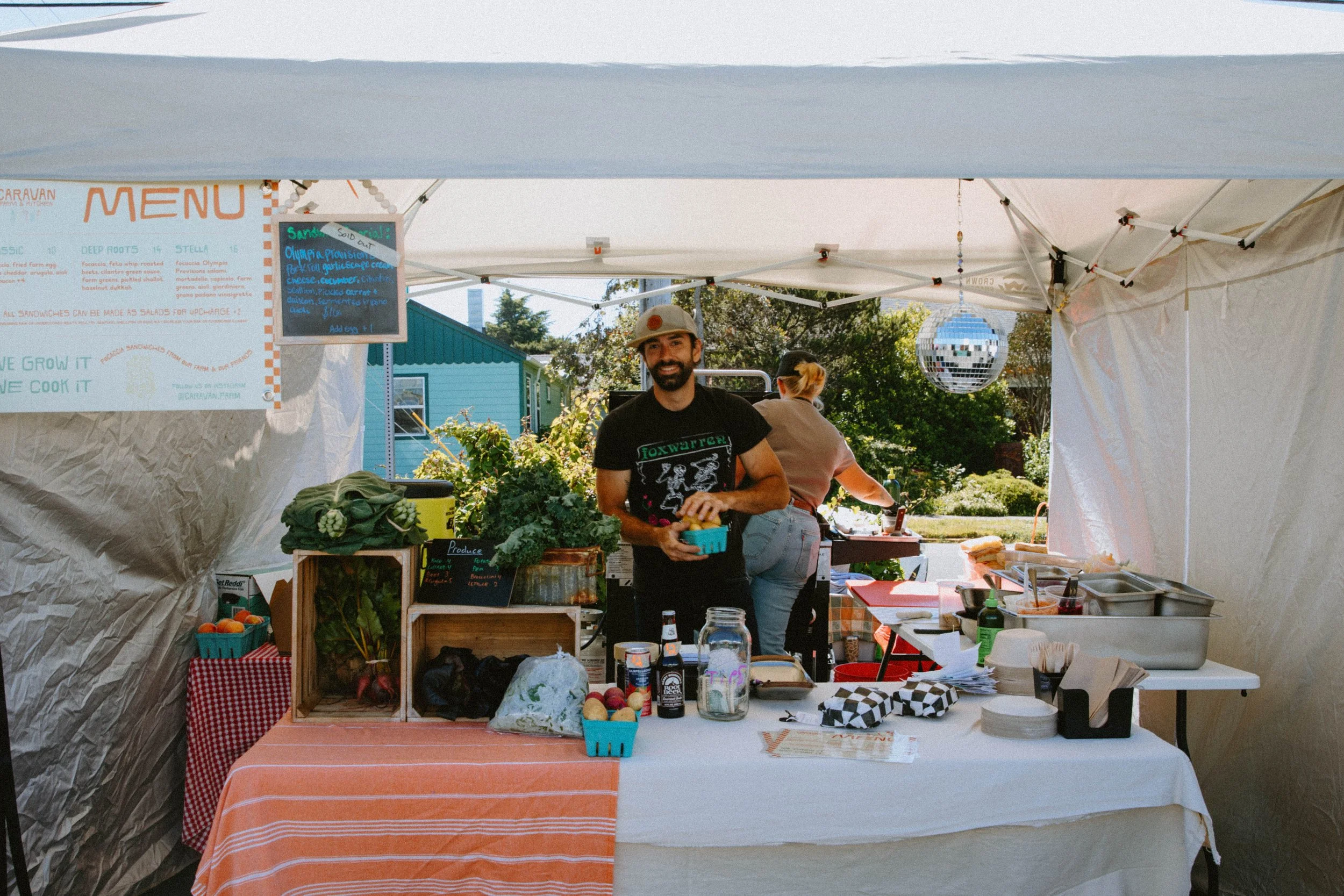 A man smiling at a farmers market booth with vegetables, eggs, and drinks on a table covered with an orange and white striped tablecloth. A woman is behind him, and there are trees and colorful buildings in the background.