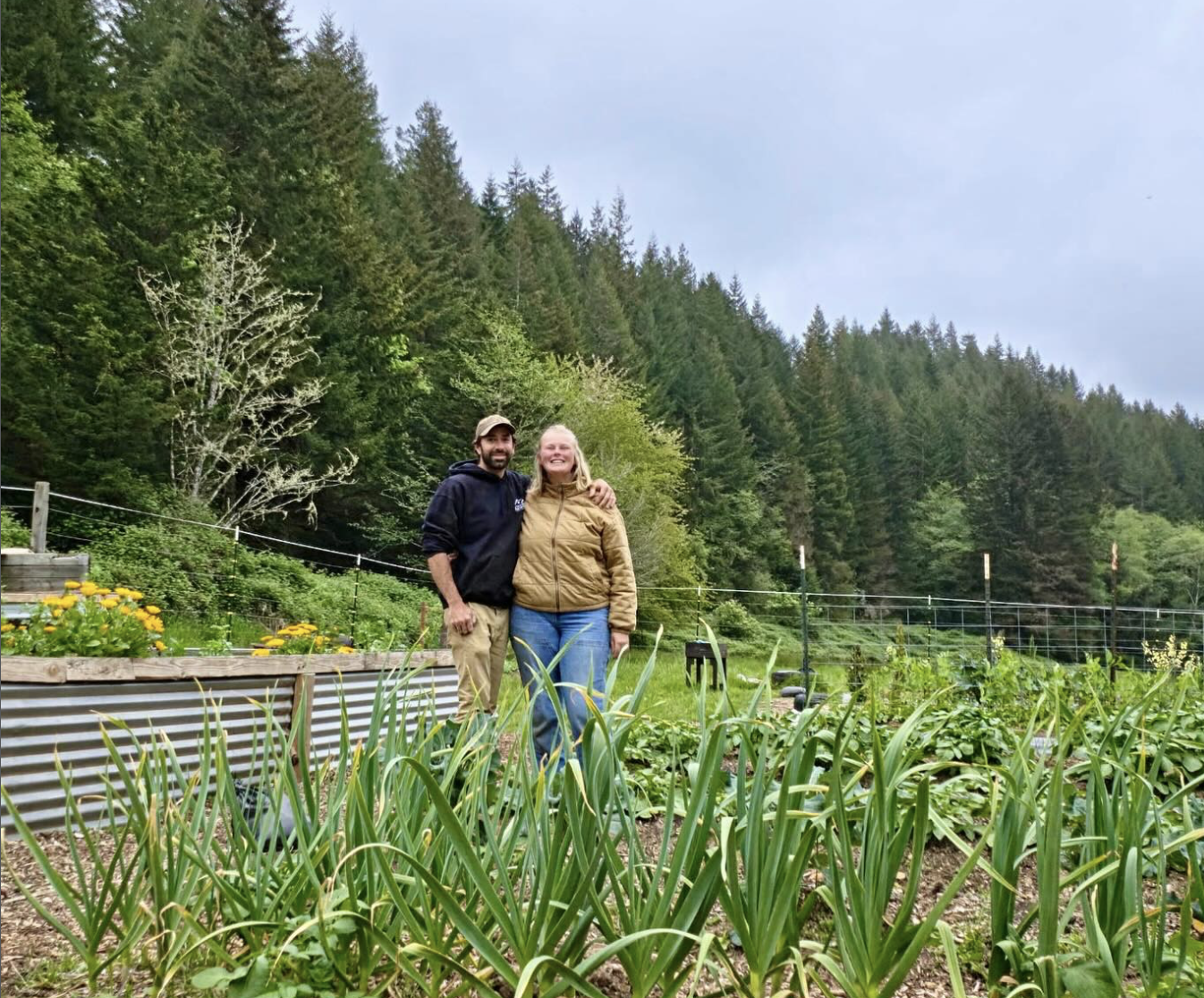 A man and woman smiling and posing in a garden with lush greenery and tall trees in the background.