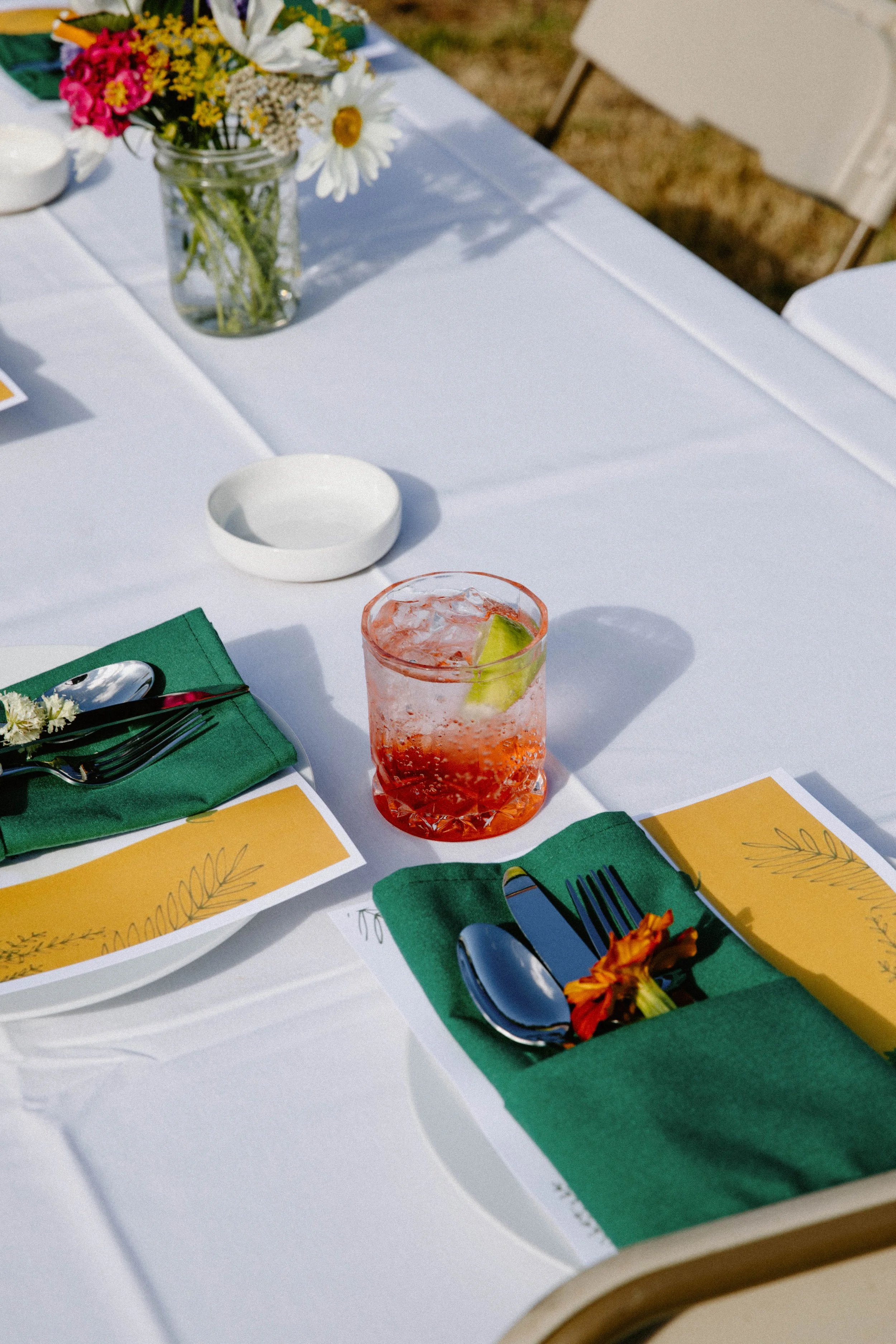 Table setting with a floral centerpiece, drinking glass with a red beverage and ice, yellow pamphlets, green napkins, and silverware on a white tablecloth.