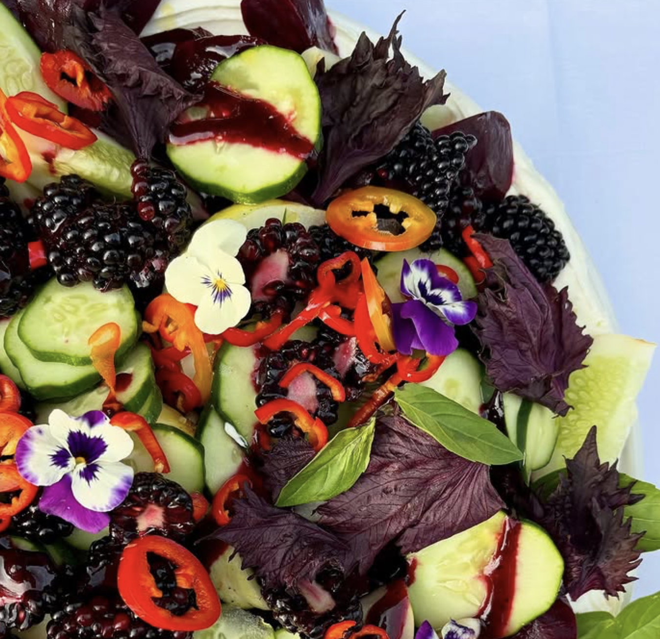 Fresh garden salad with sliced cucumbers, blackberries, red and yellow chili peppers, edible flowers, and green and purple herbs.