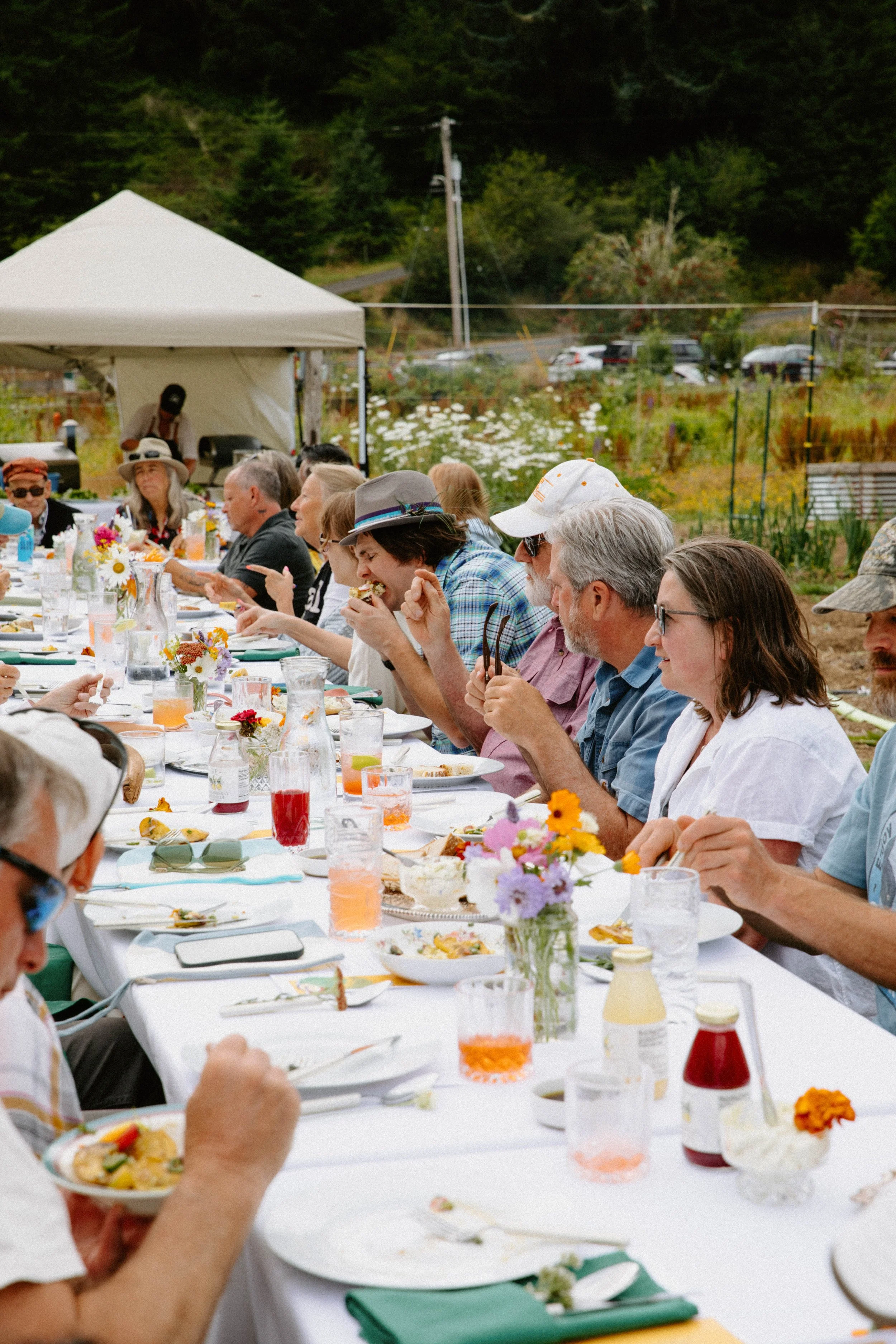 People seated at a long outdoor banquet table set with flowers, drinks, and plates of food, enjoying a meal together in a garden setting during daytime.