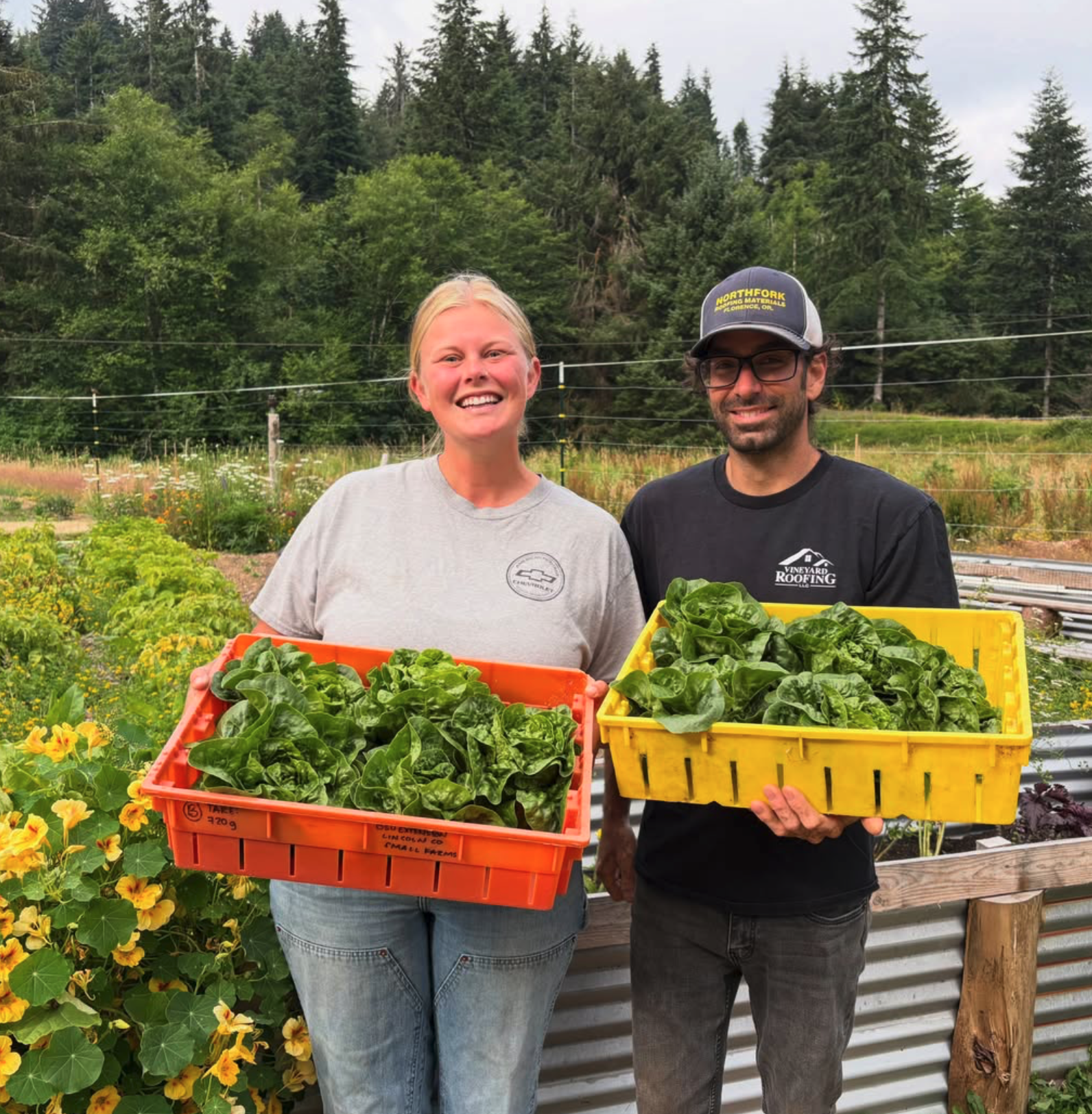 Two people smiling and holding large containers of fresh lettuce in a garden with flowers and trees in the background.