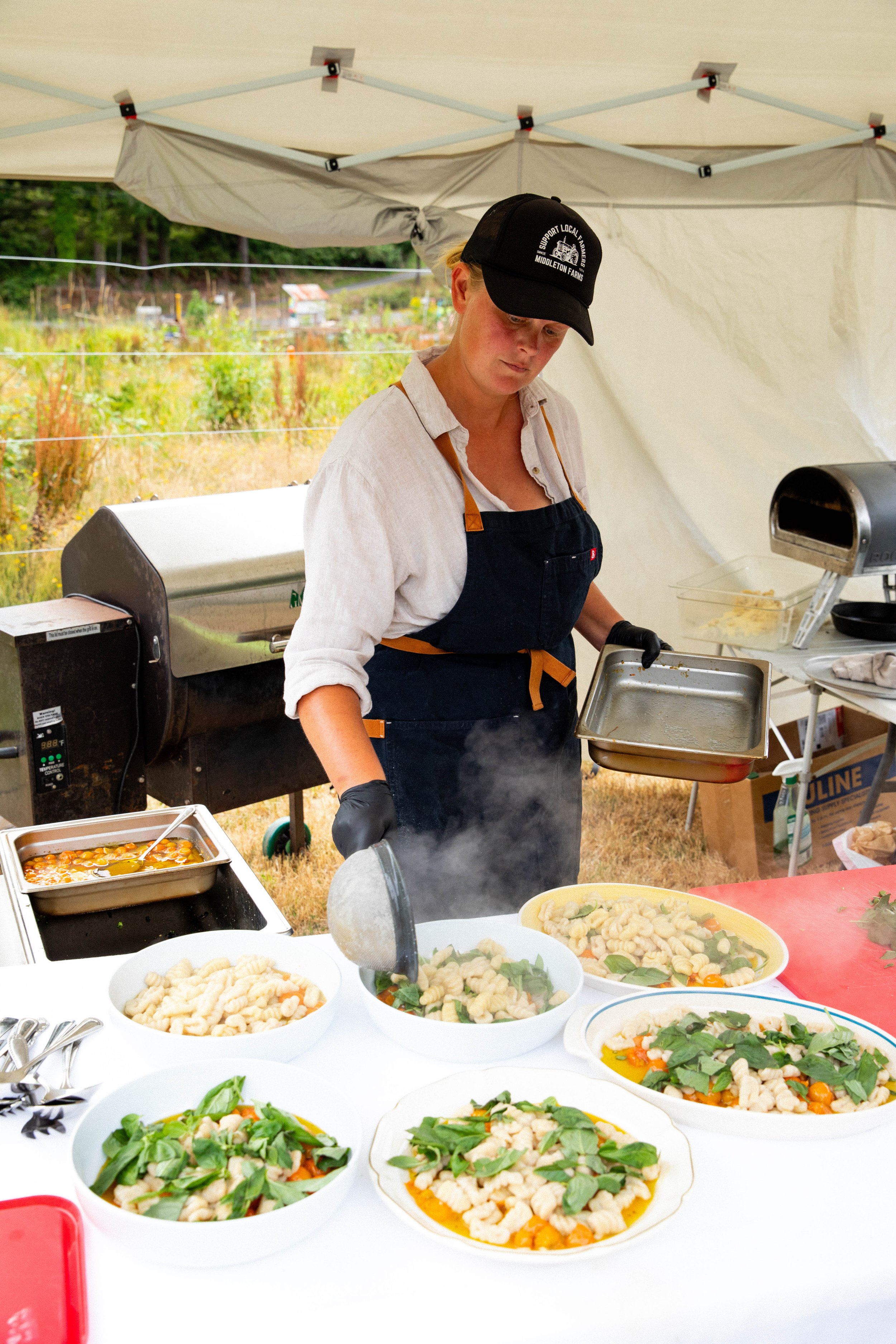 A woman wearing a black cap and apron serving food at an outdoor buffet with bowls of pasta and salads on a table.