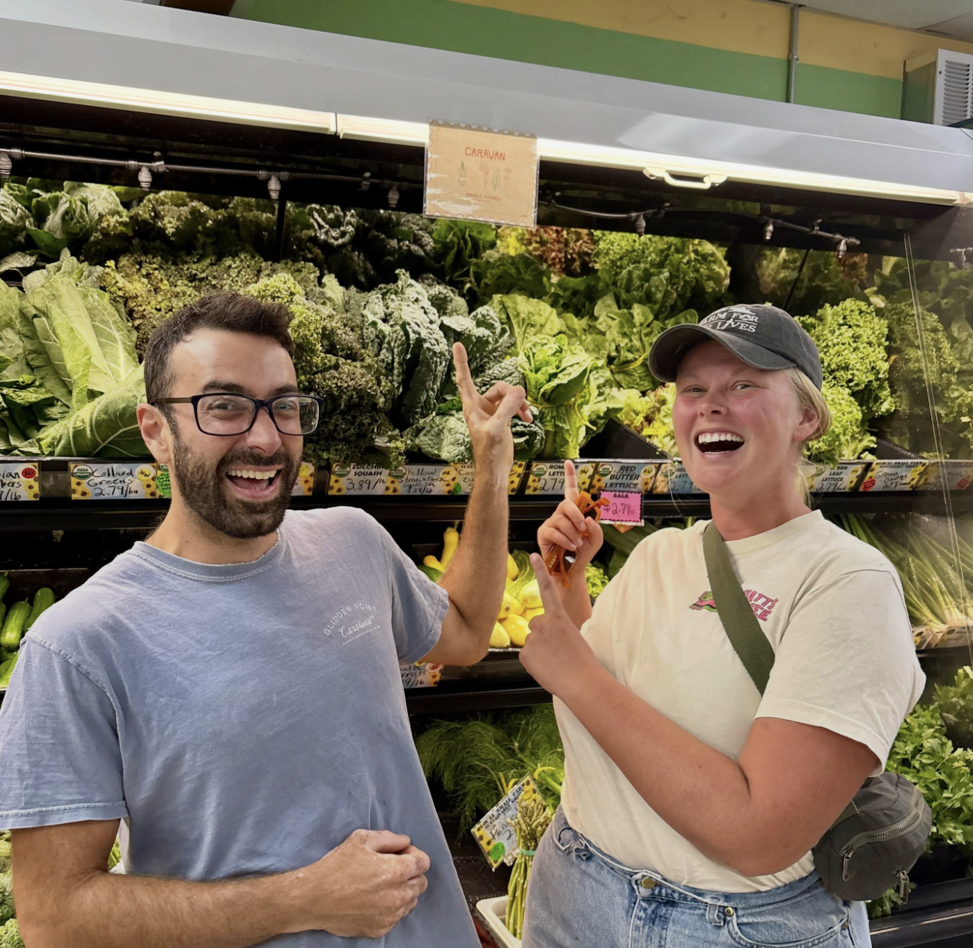 Two smiling people, a man with glasses and a woman wearing a cap, standing in front of a display of leafy greens at a grocery store. They are pointing at each other and appear to be happy.