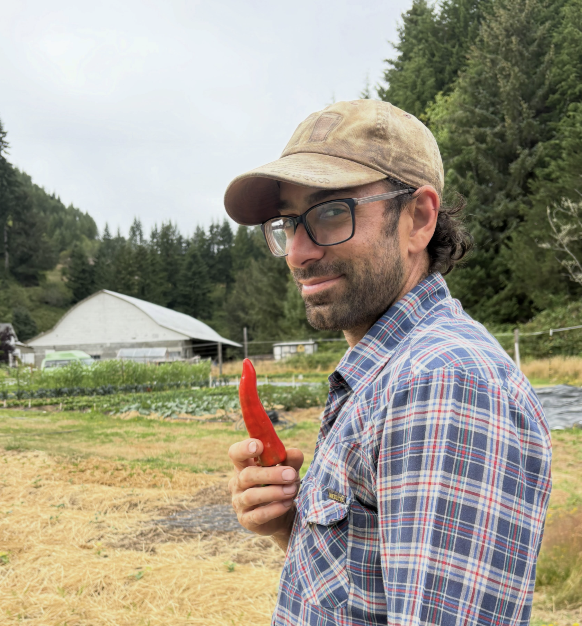 A man with glasses and a plaid shirt holding a red chili pepper outdoors on a farm, with a barn and trees in the background.