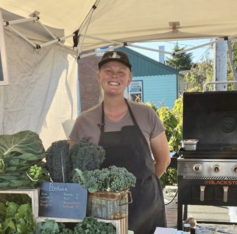 Smiling woman standing behind a produce stand with broccoli and other vegetables at an outdoor market.