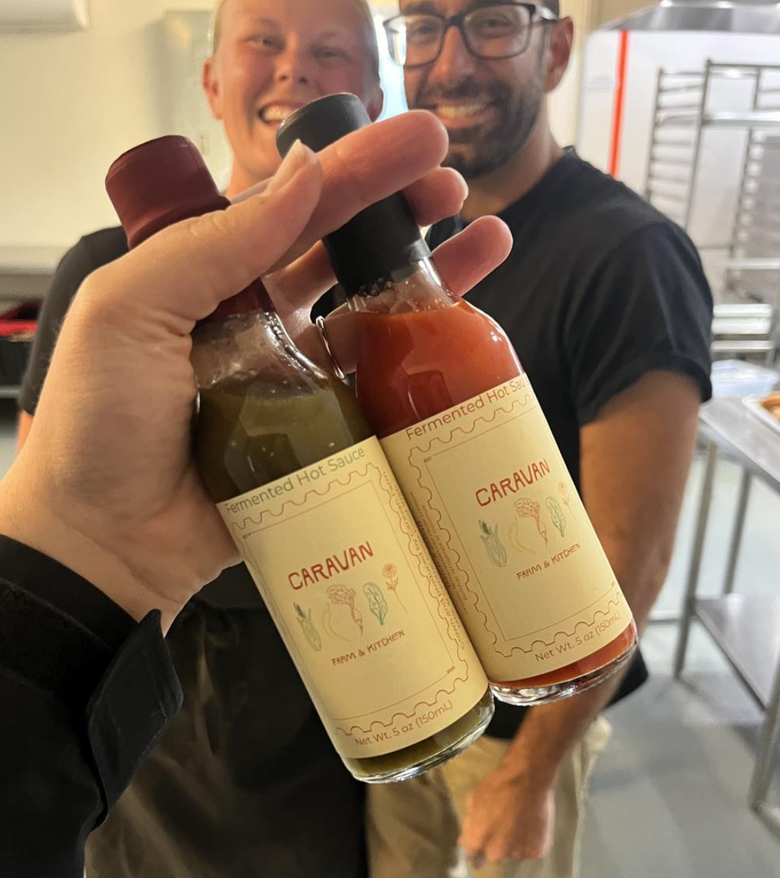 Two bottles of hot sauce labeled 'Caruvan' held in front of two smiling men in a kitchen or restaurant setting.