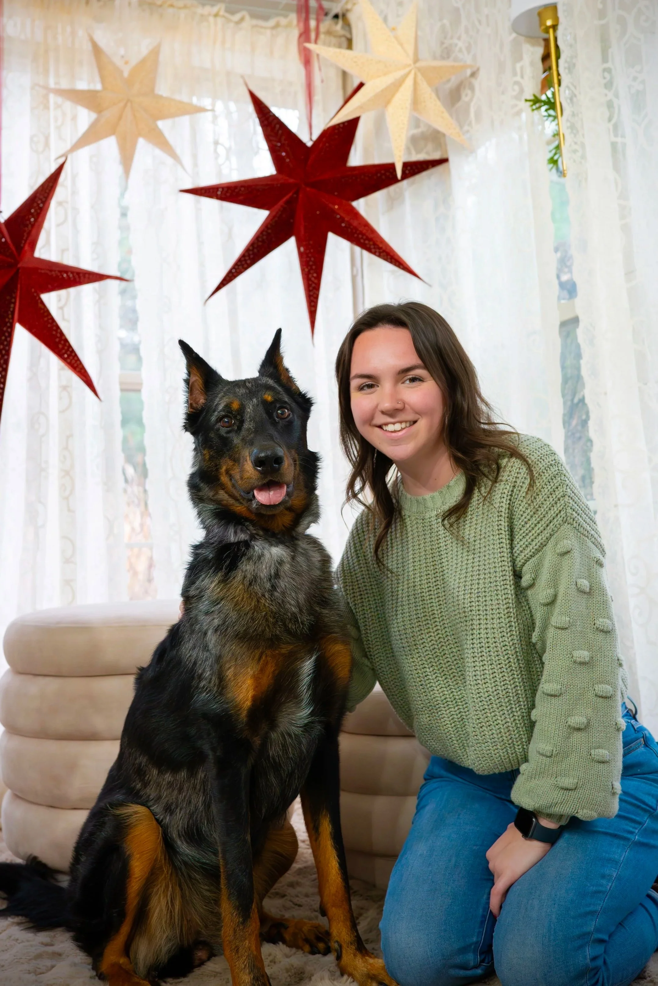 A young woman with brown hair, wearing a green sweater and blue jeans, is kneeling on the floor next to a large black and tan dog with a coat that has speckled black and gray markings. The dog is sitting inside a room decorated for Christmas with red and cream paper star lanterns hanging from the ceiling and white lace curtains covering the windows in the background.