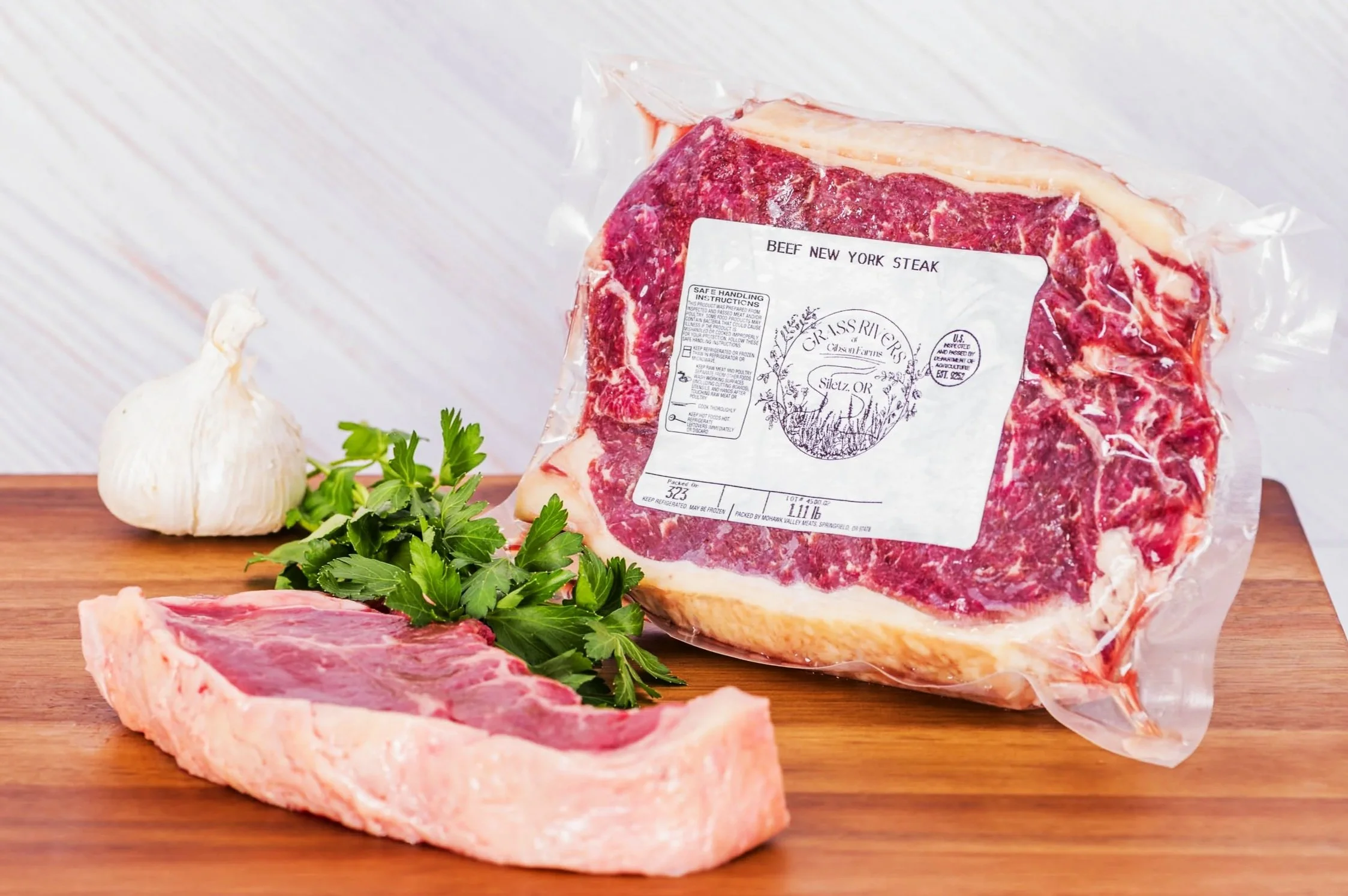 Package of beef New York steak, garlic, parsley, and raw beef cut on a wooden surface.