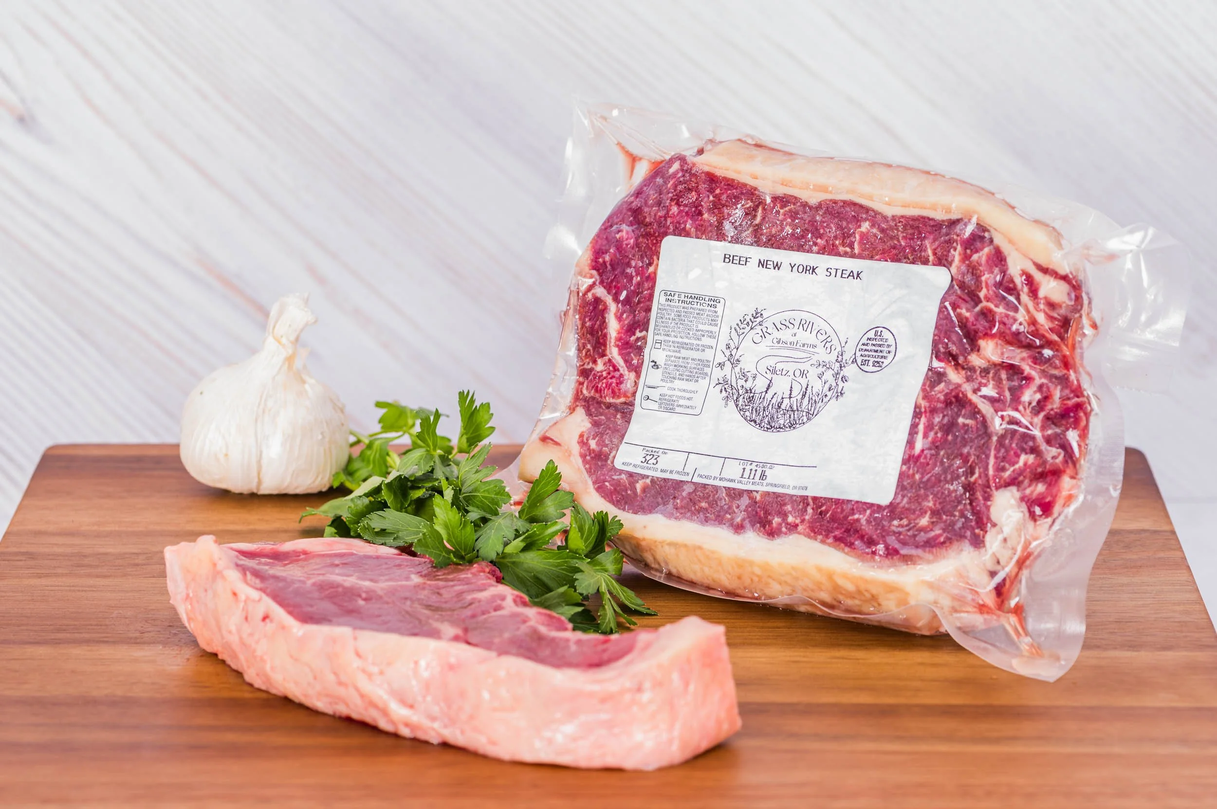 A vacuum-sealed package of beef New York steak on a wooden surface with garlic, fresh parsley, and raw steak slices placed nearby.