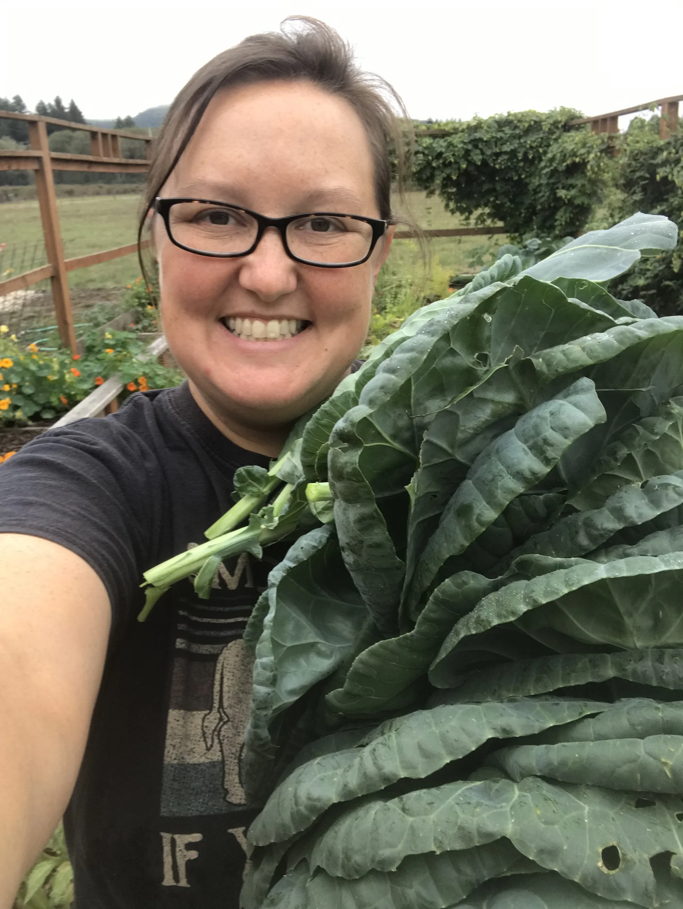 A person with short brown hair, glasses, and a black shirt smiling in a garden, holding large green leafy vegetables.