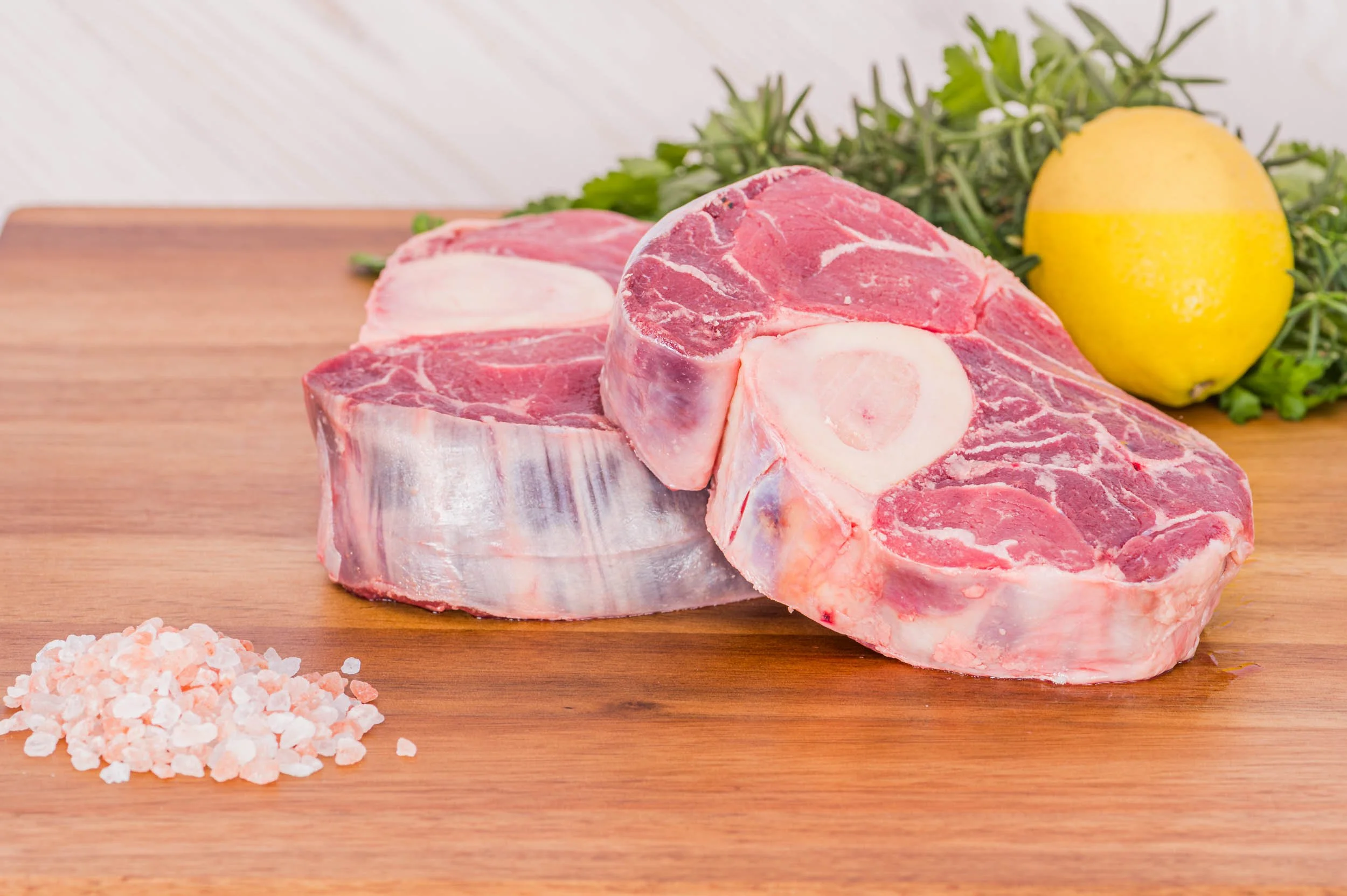 Cut raw beef shank on wooden cutting board with pink Himalayan salt, a lemon, and green herbs in the background.