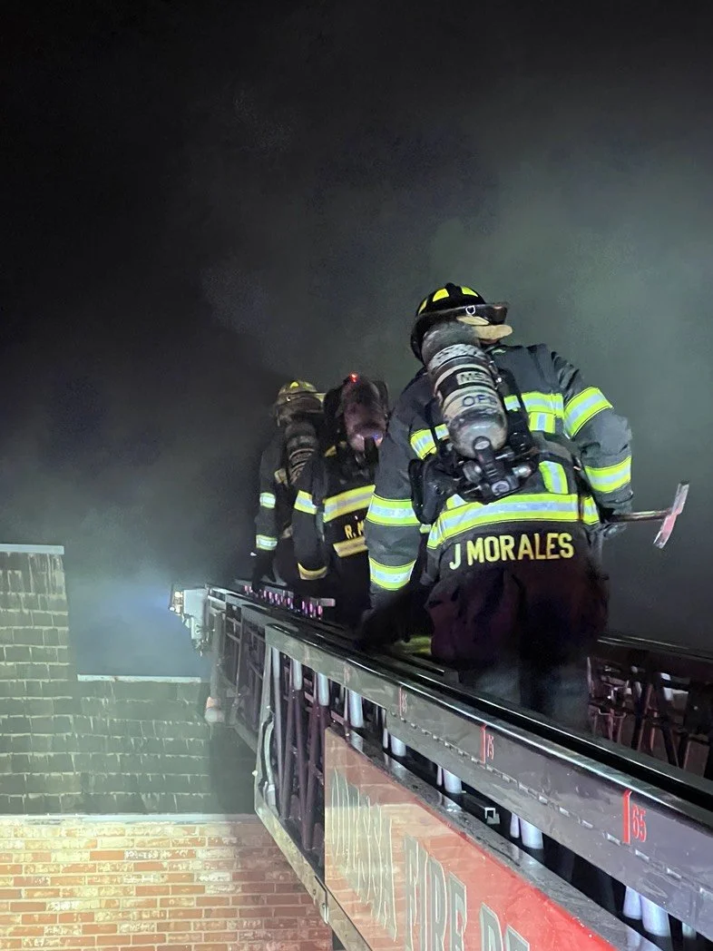 Firefighters on an upper fire truck ladder responding to a fire at night, with smoke in the background.