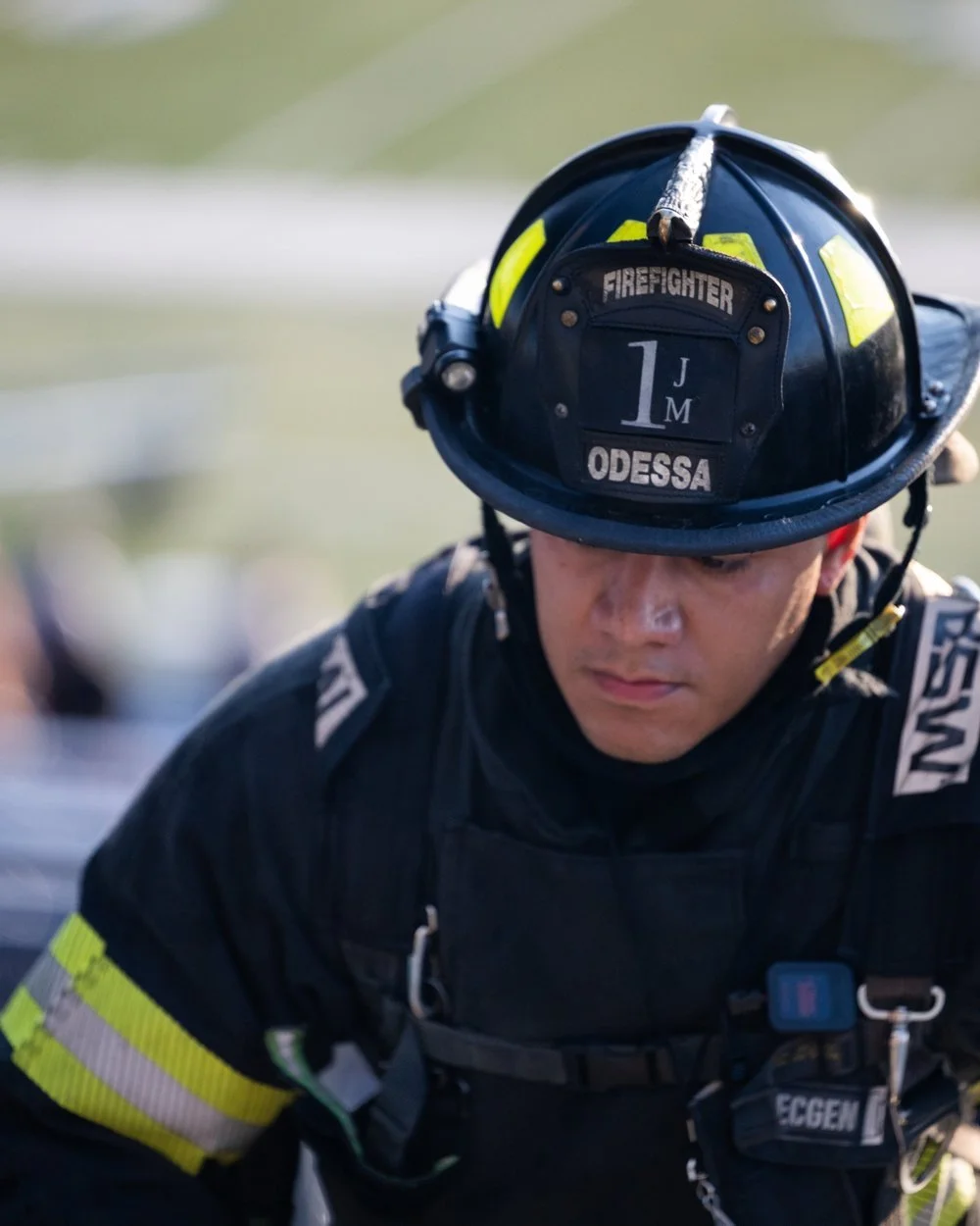 A firefighter in black uniform with reflective stripes, wearing a black helmet with yellow accents, labeled 'FIRE FIGHTER,' 'ODESSA,' and the number 1. The firefighter is looking down with a serious expression.