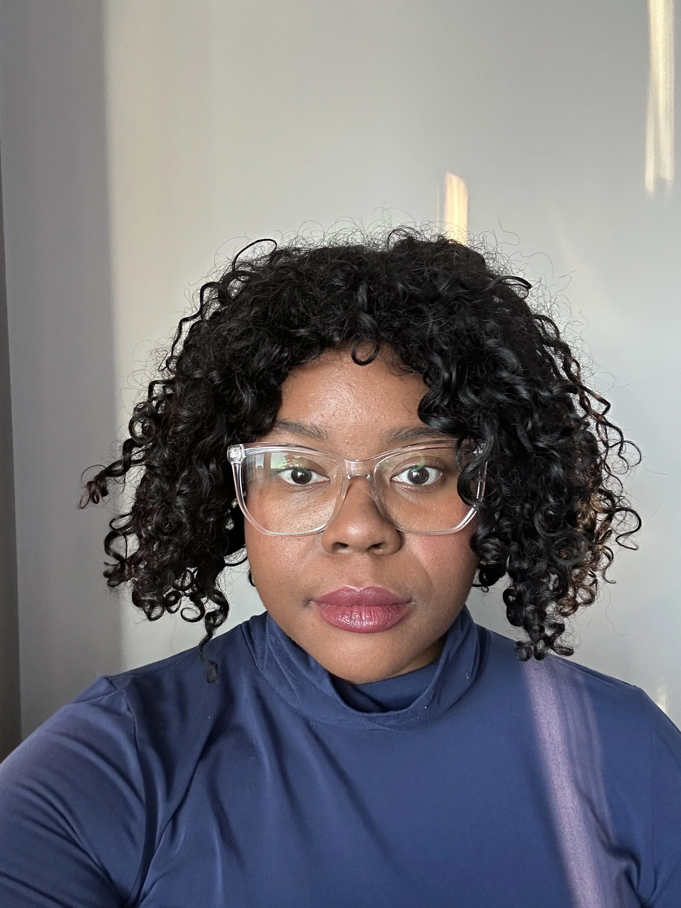 A woman with curly black hair and glasses taking a selfie, wearing a navy blue top, against a plain light-colored background.