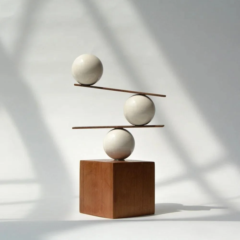 Balancing sculpture with three white spheres stacked on a wooden base and two wooden sticks connecting the spheres.