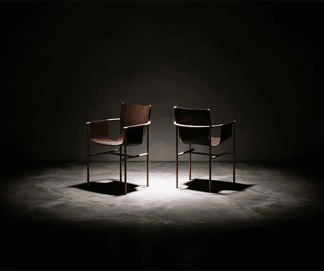Two chairs with armrests facing each other in a dark, empty room with a spotlight overhead.