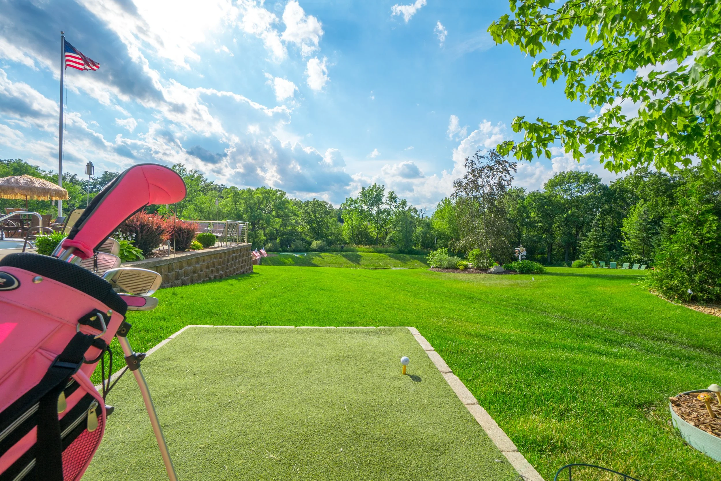 A golf course with a pink golf bag and clubs in the foreground, a tee box with a golf ball, lush green trees, and a partly cloudy sky with an American flag in the background.