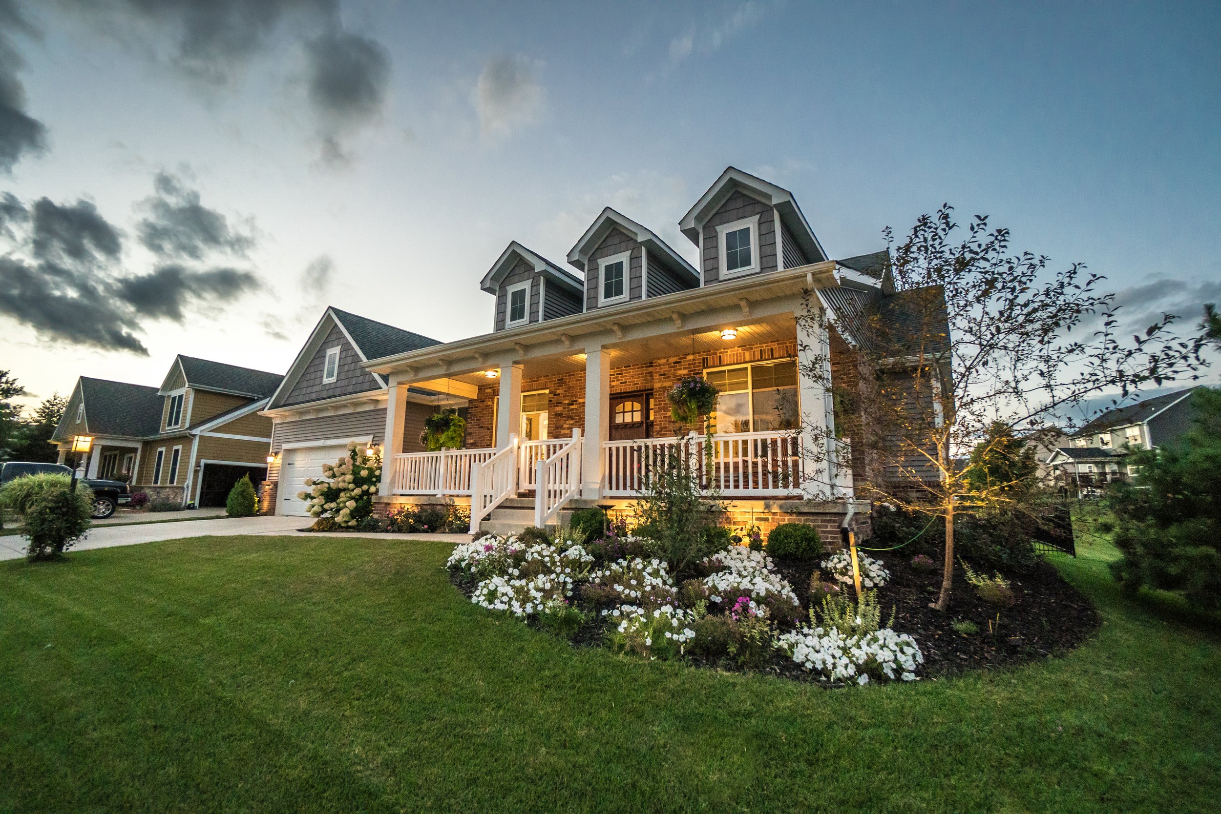 A two-story suburban house with a brick and siding exterior, illuminated porch lights, a white porch railing with hanging flowers, and a well-manicured lawn with flower beds. The sky is partly cloudy at dusk.