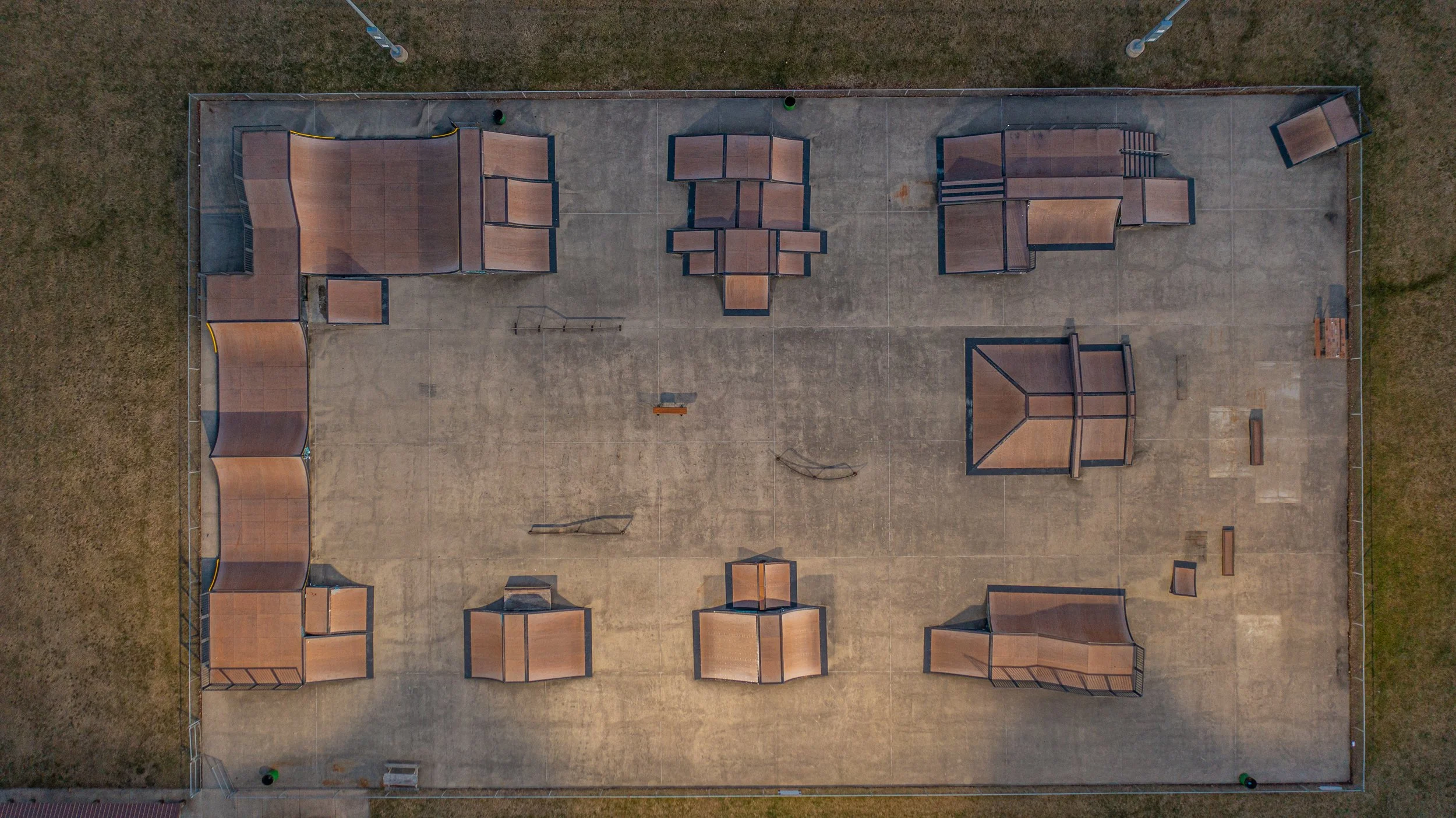 Aerial view of a skatepark with various ramps, rails, and platforms on a concrete surface, enclosed by a chain-link fence with grassy areas outside.