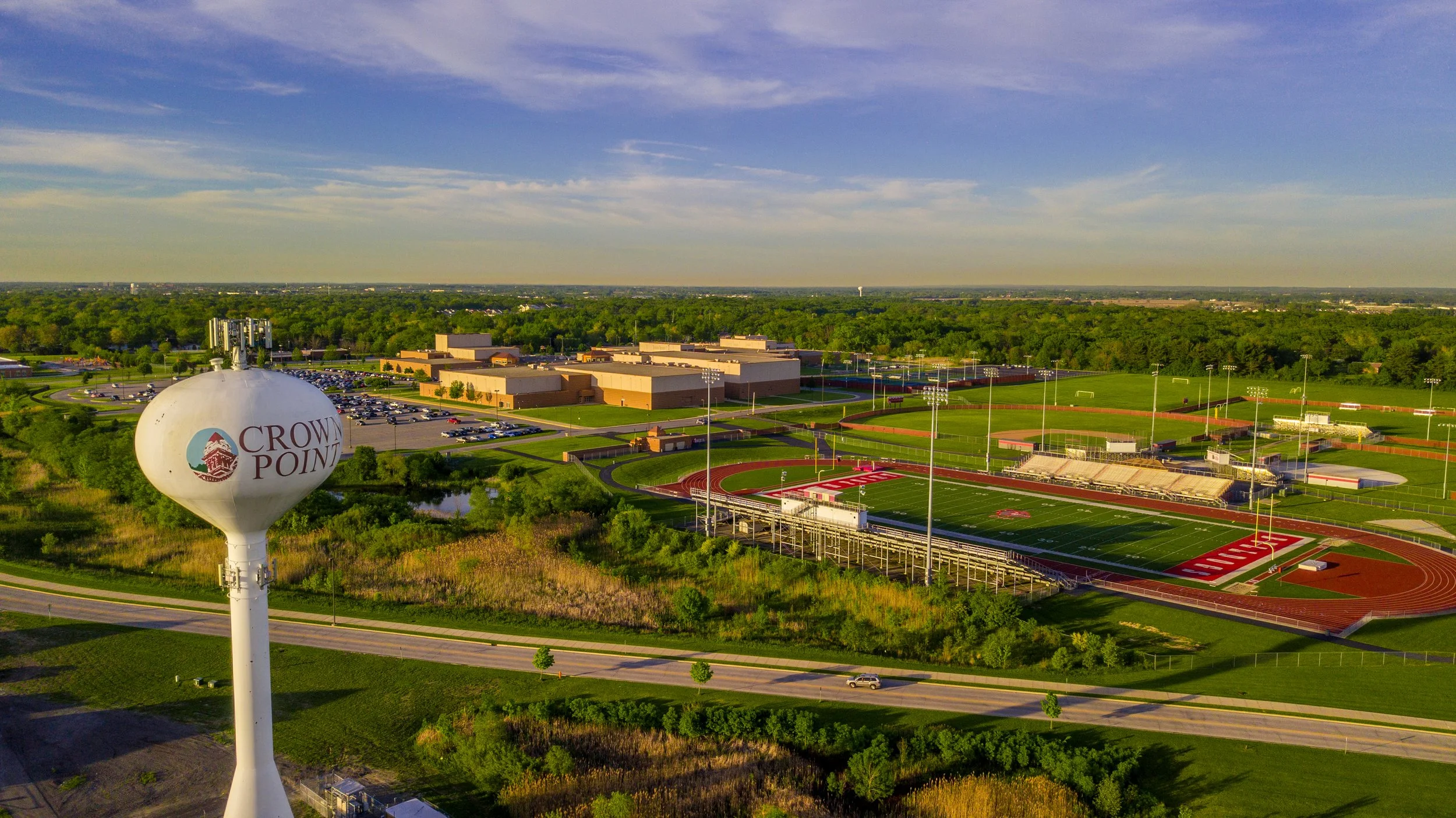 Aerial view of a high school campus with athletic fields, parking lot, and a water tower labeled 'Crown Point' under a partly cloudy sky.