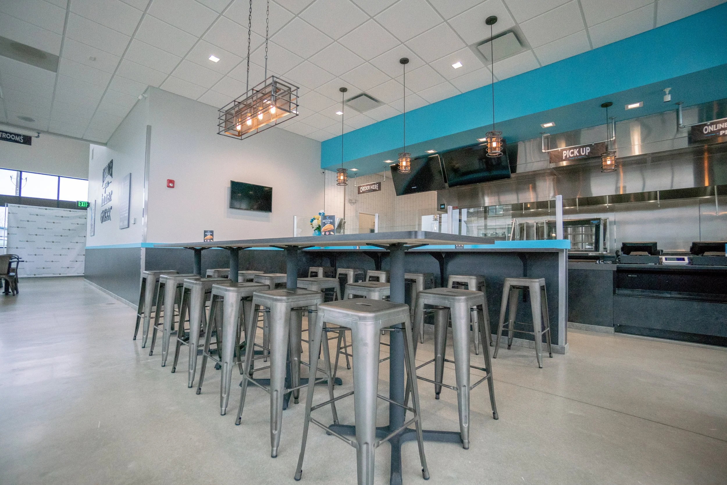 Interior of a modern fast-food restaurant with a long communal table surrounded by metal stools, hanging pendant lights, a television screen, and signs indicating order and pick-up areas.