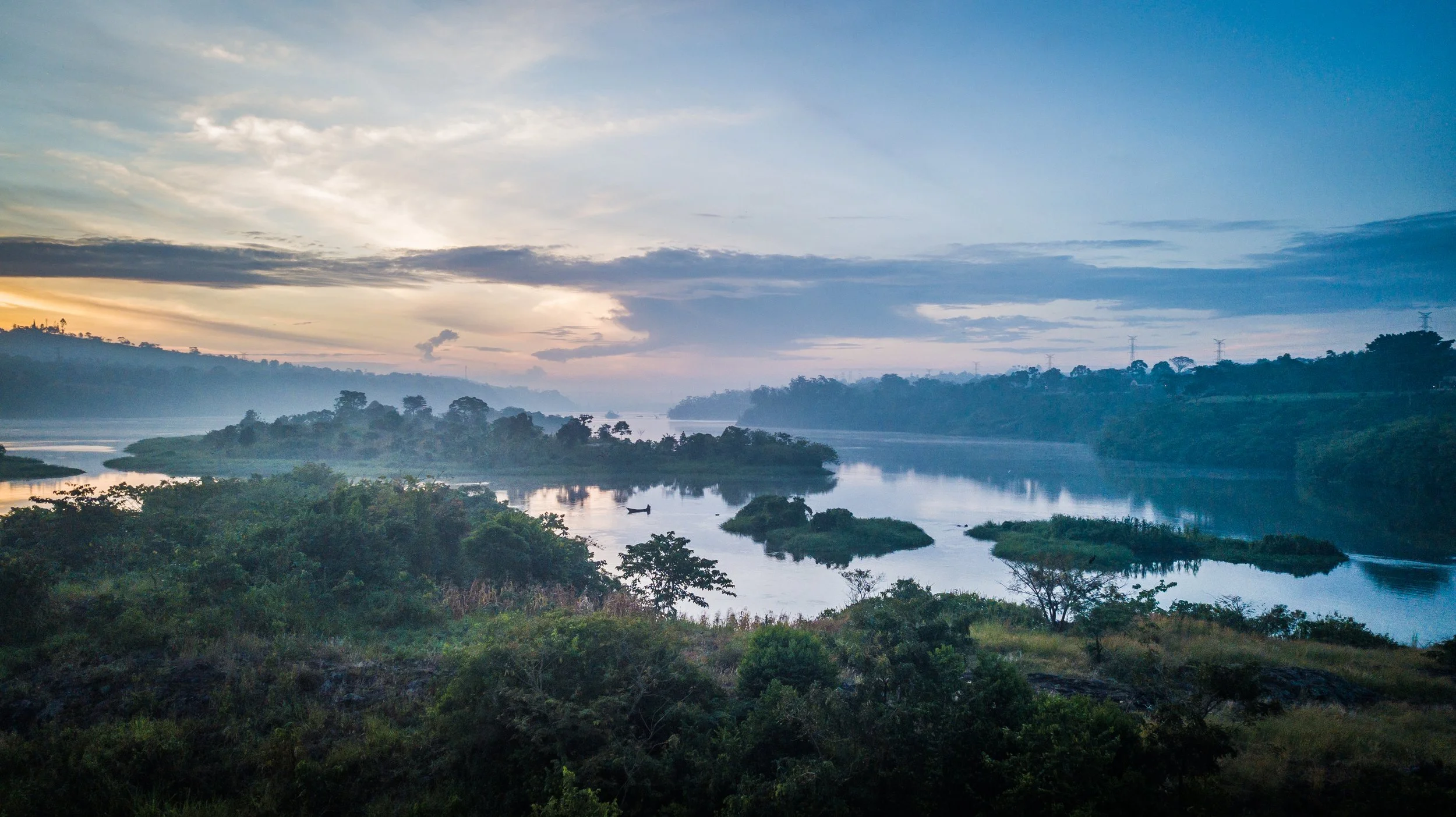 A river flowing through a lush green landscape with trees and shrubs, under a partly cloudy sky during dawn or dusk.