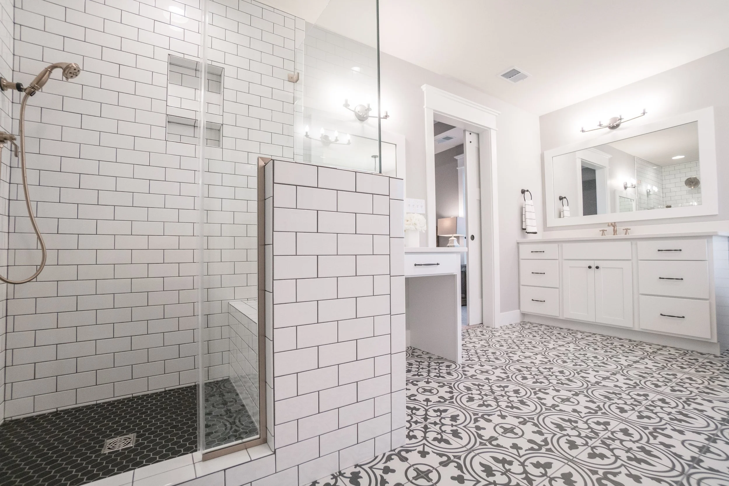 Modern bathroom with a glass-enclosed shower featuring white subway tiles and black hexagon tiles on the shower floor. The bathroom has a large mirror above a white vanity with black handles, decorated with striped towels and a small vase with flower