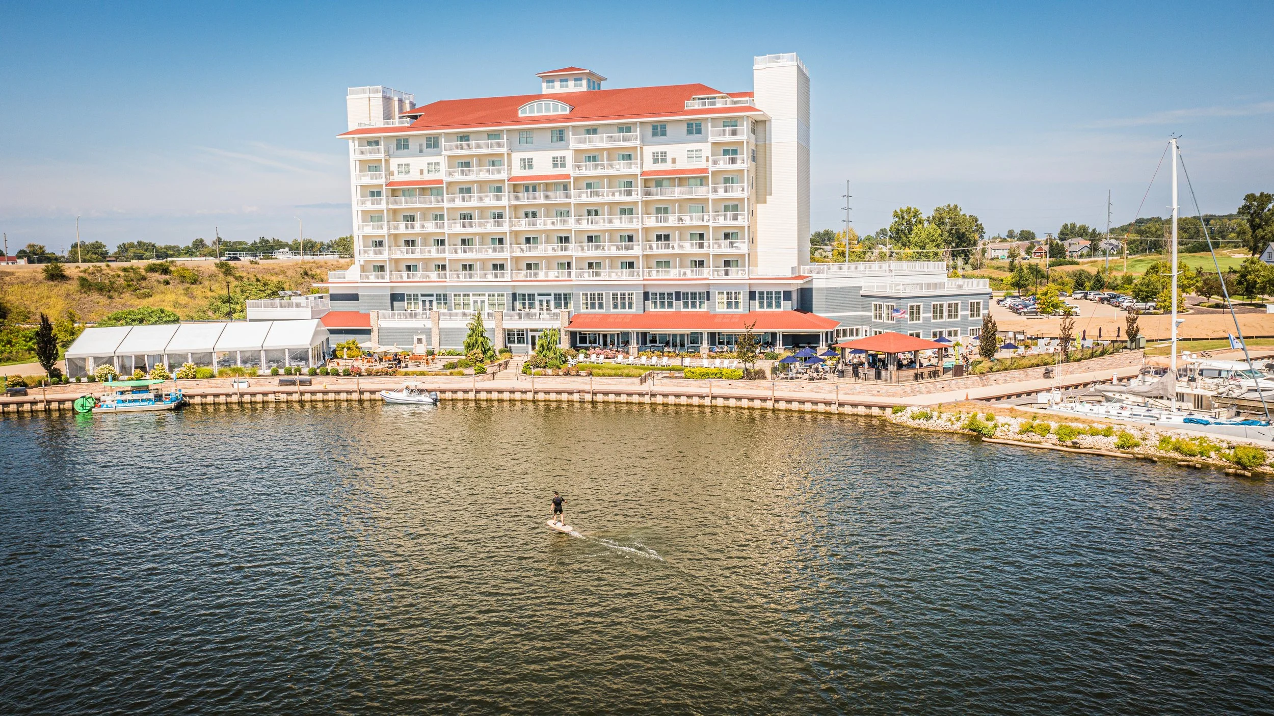 Large multi-story waterfront building with balconies, outdoor seating area, and boats docked nearby, situated beside a body of water under a clear blue sky.