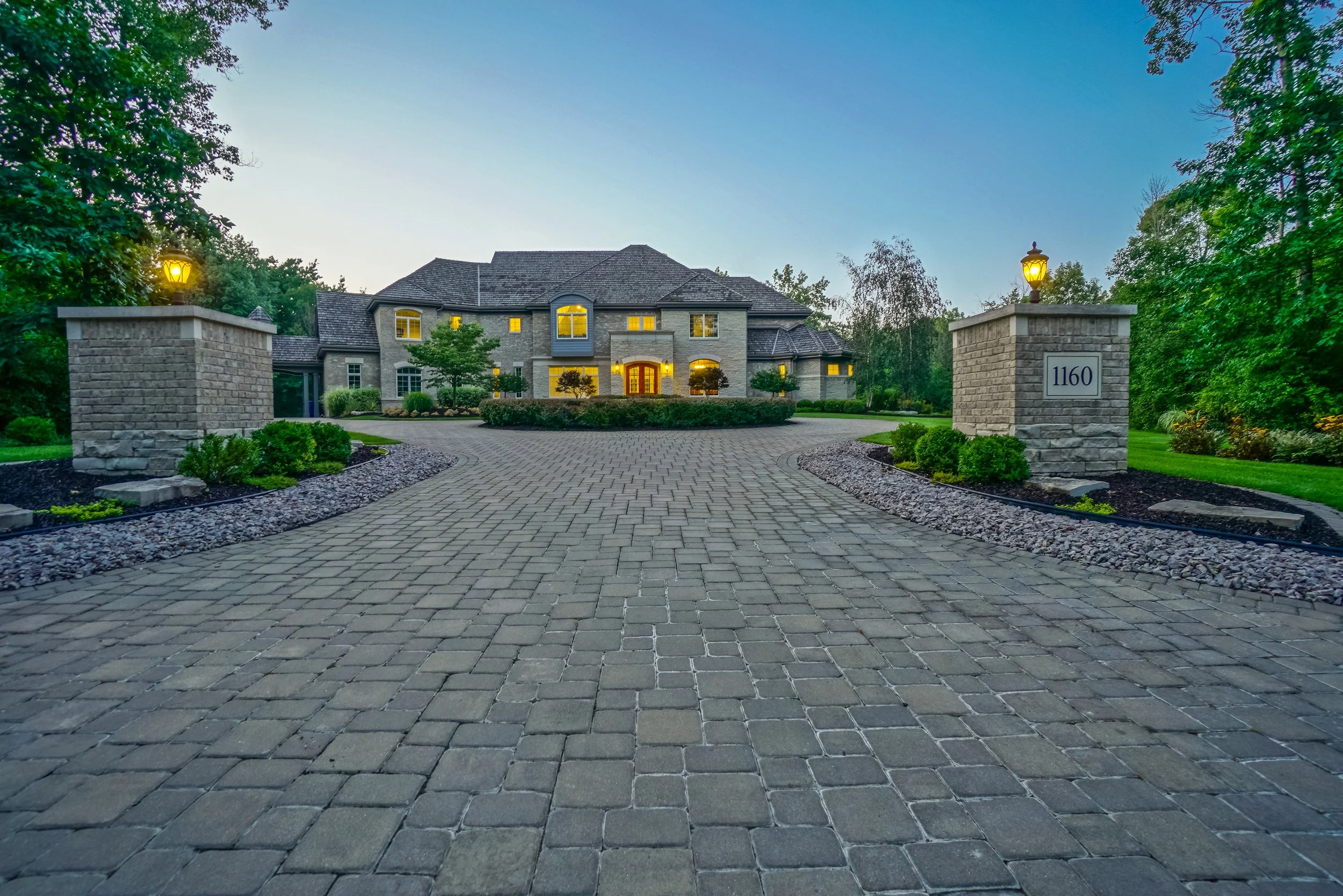 A large, elegant house with multiple windows and a brick facade, surrounded by well-maintained landscaping and trees, with a cobblestone driveway leading up to the front entrance. The house is illuminated from inside, with lamps glowing on the brick pillars at the entrance.
