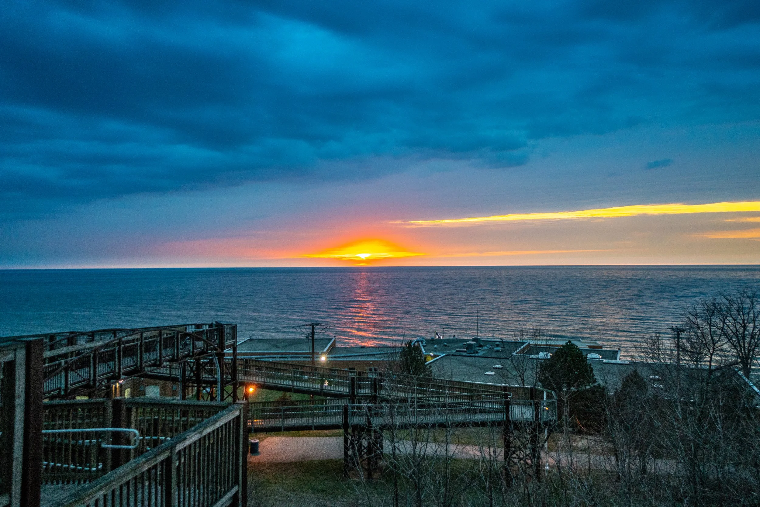 Sunset over the ocean with a partly cloudy sky, view from a seaside structure with wooden walkways and rooftops in the foreground.