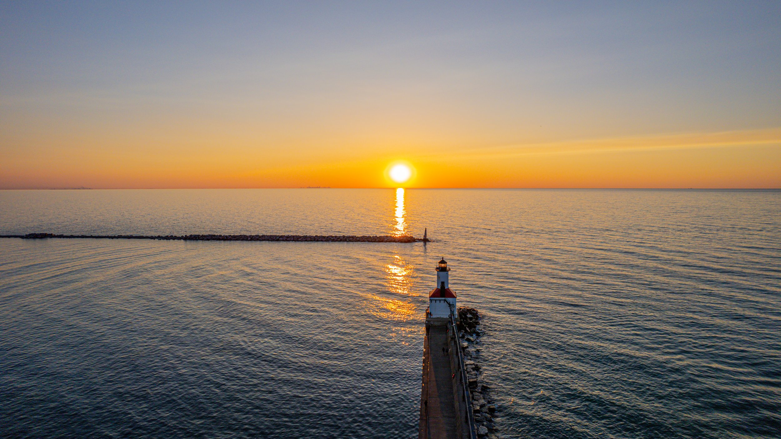 A lighthouse is situated at the end of a pier extending into a large body of water. The sun is setting on the horizon, casting a golden reflection on the water. The sky is clear with a gradient from blue to orange.