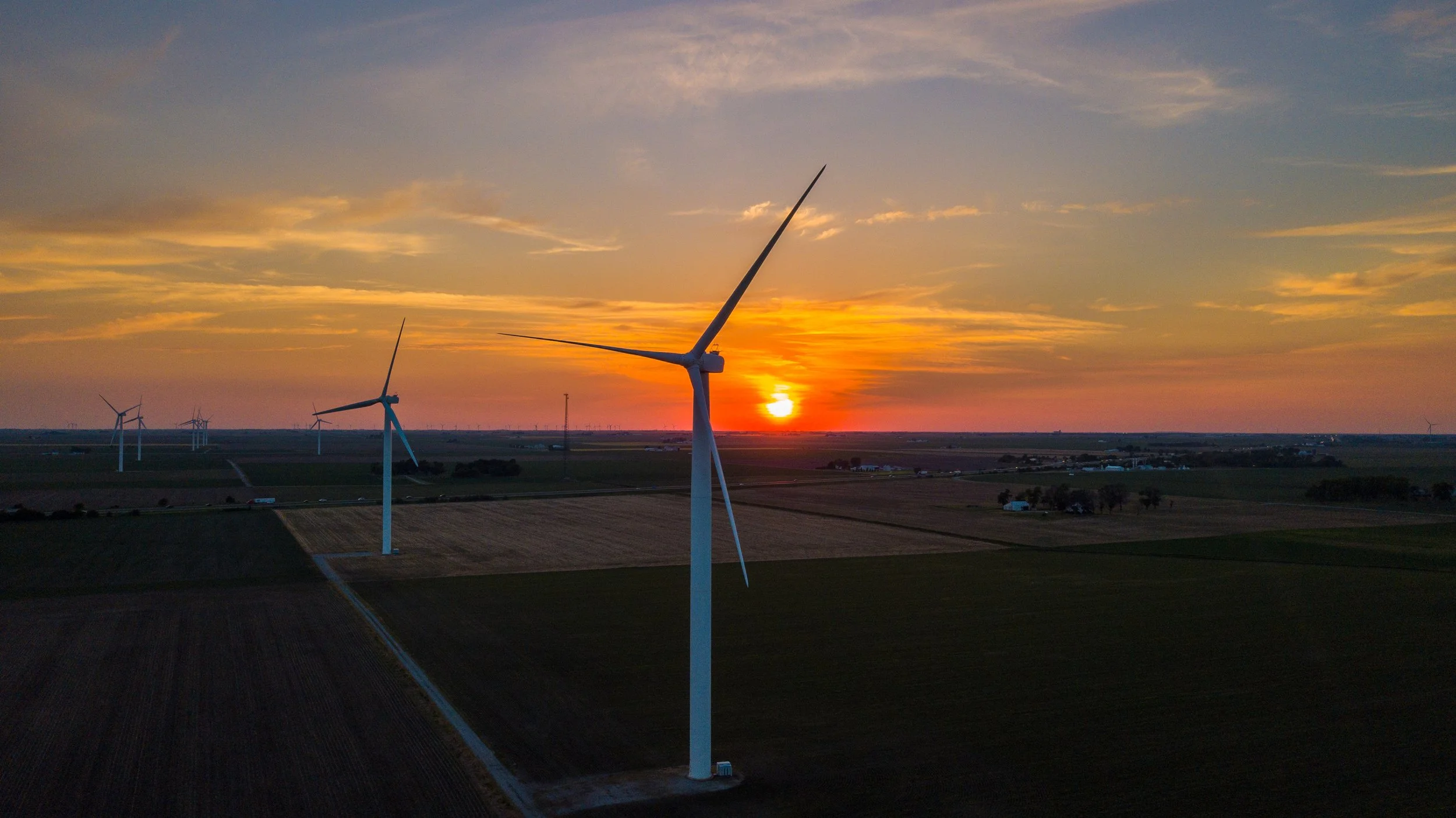 A wind farm at sunset with several wind turbines on farmland under a sky with orange, yellow, and blue clouds.