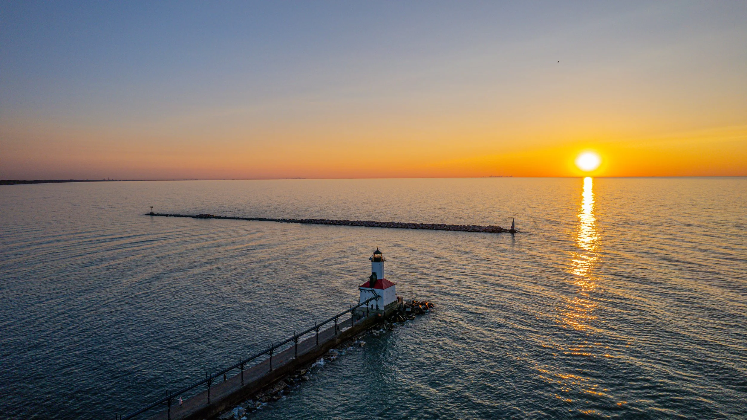 Sunset over the water with a lighthouse on a pier and a breakwater, calm ocean, sky transitioning from blue to orange