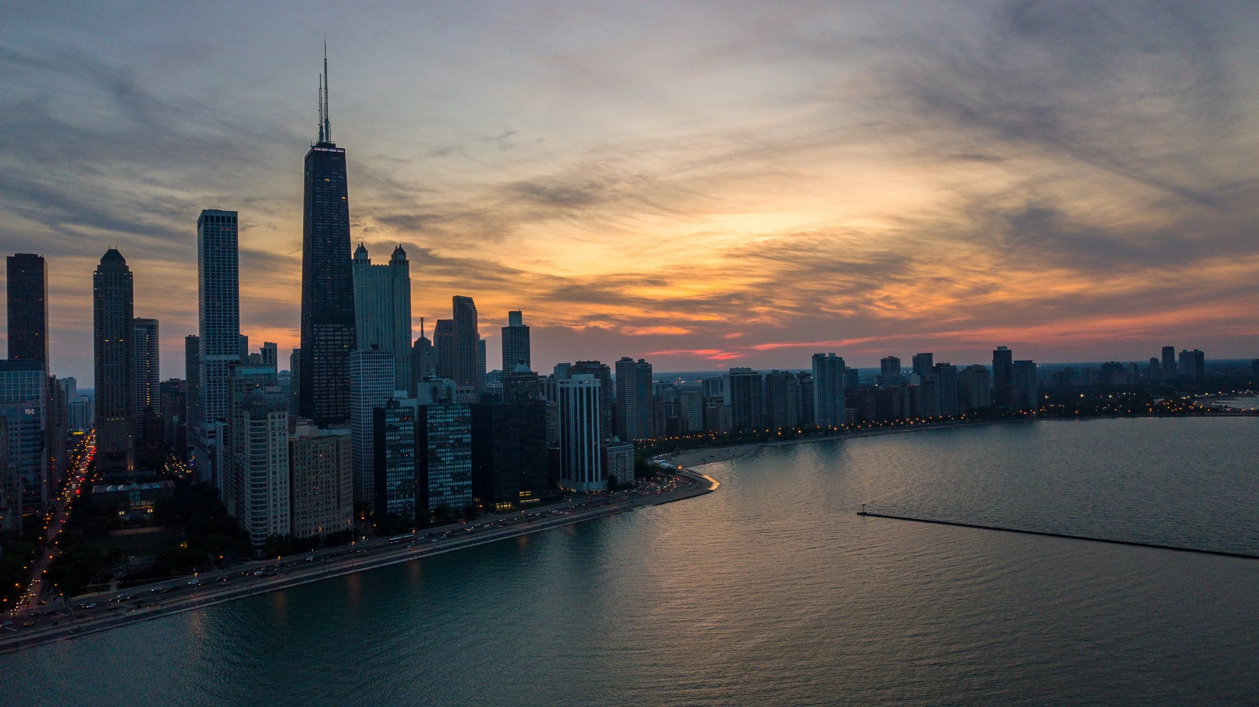 A city skyline at sunset with tall buildings, including a prominent skyscraper, along a river, with a road running parallel to the water and city lights beginning to turn on.