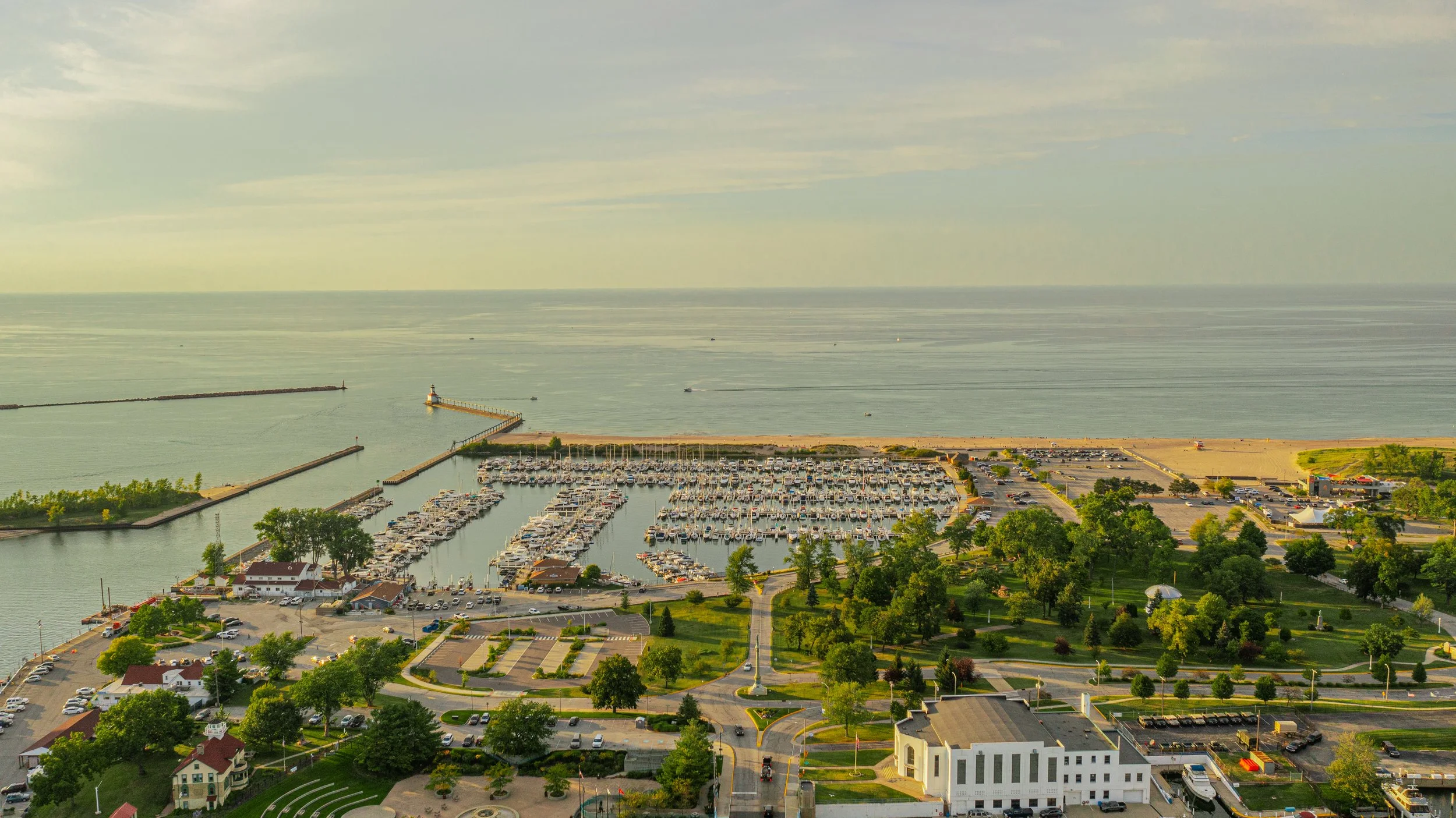 Aerial view of a marina with boats, a parking lot, and surrounding greenery overlooking a large body of water with a small lighthouse and breakwater.