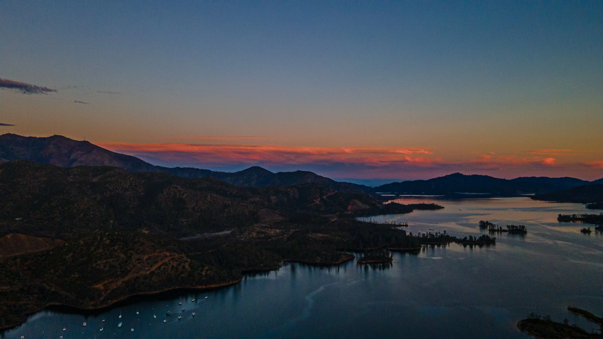 Scenic view of a lake with boats, surrounded by mountains, under a colorful sunset sky with pink and orange clouds.