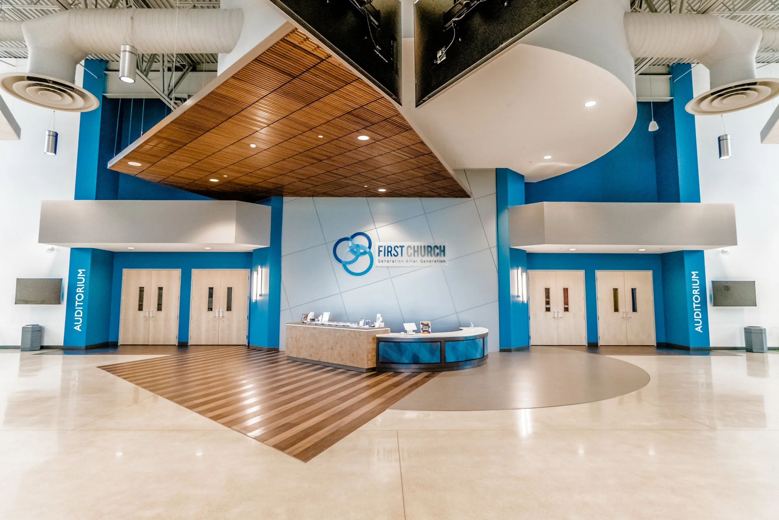 The entrance to a church auditorium with a reception desk in the center and doors on either side. The church's logo and name are displayed on the wall behind the desk, which is decorated in blue and white with wooden accents and modern lighting.