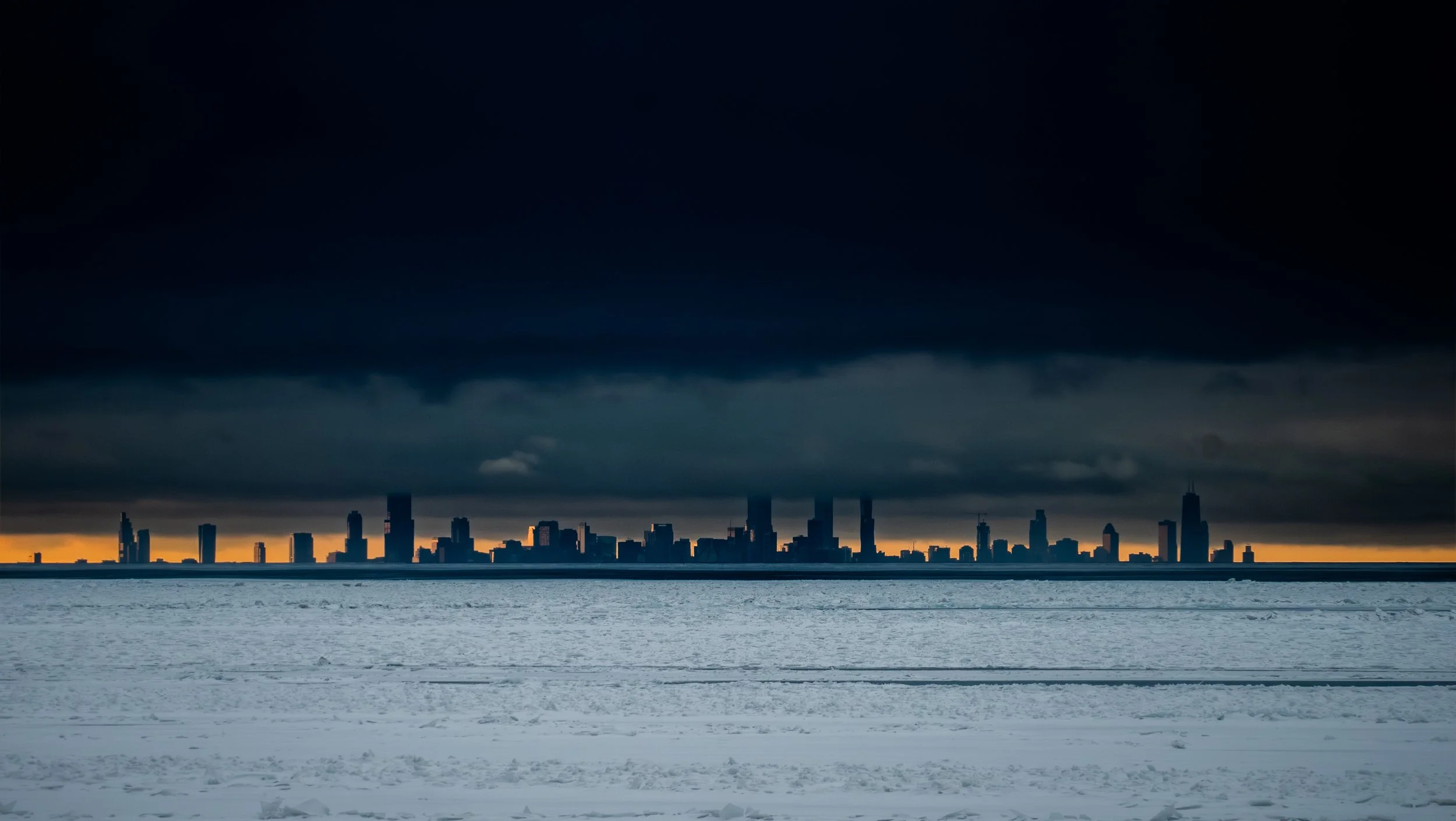 Dark clouds over a city skyline at sunset, with snow-covered ground in the foreground.