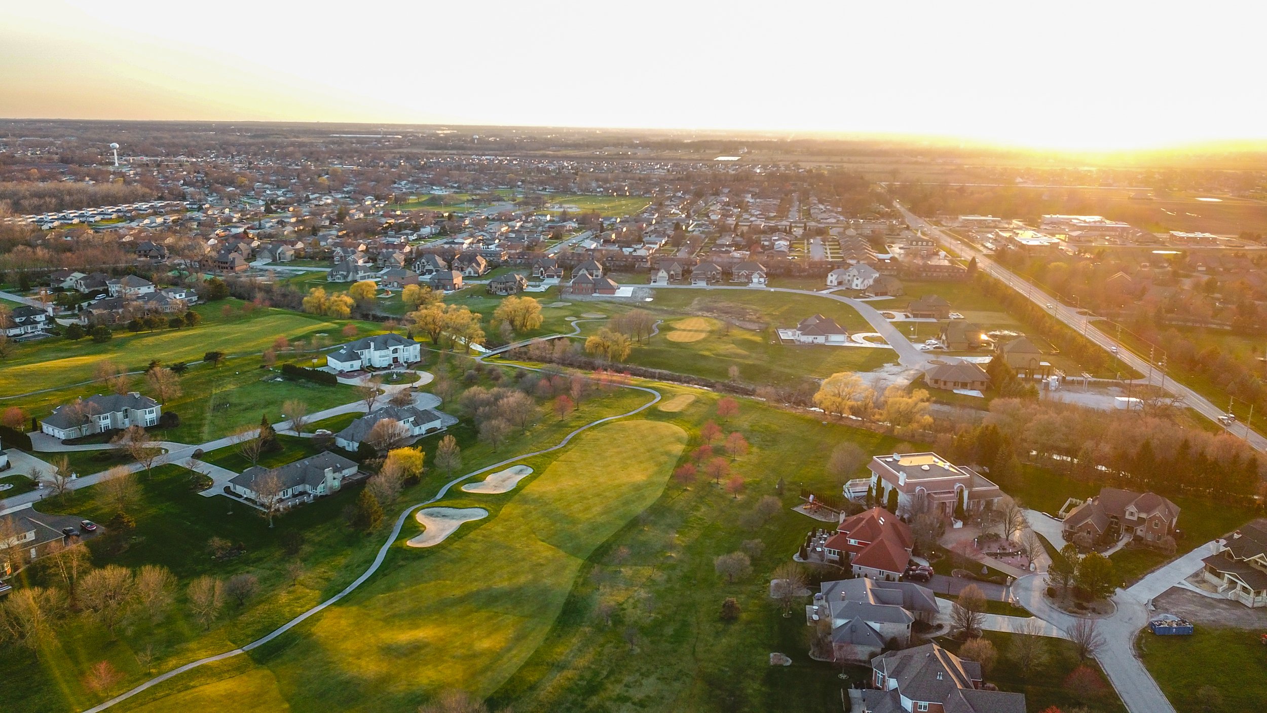 Aerial view of a suburban neighborhood with a golf course, houses, and green spaces during sunset.