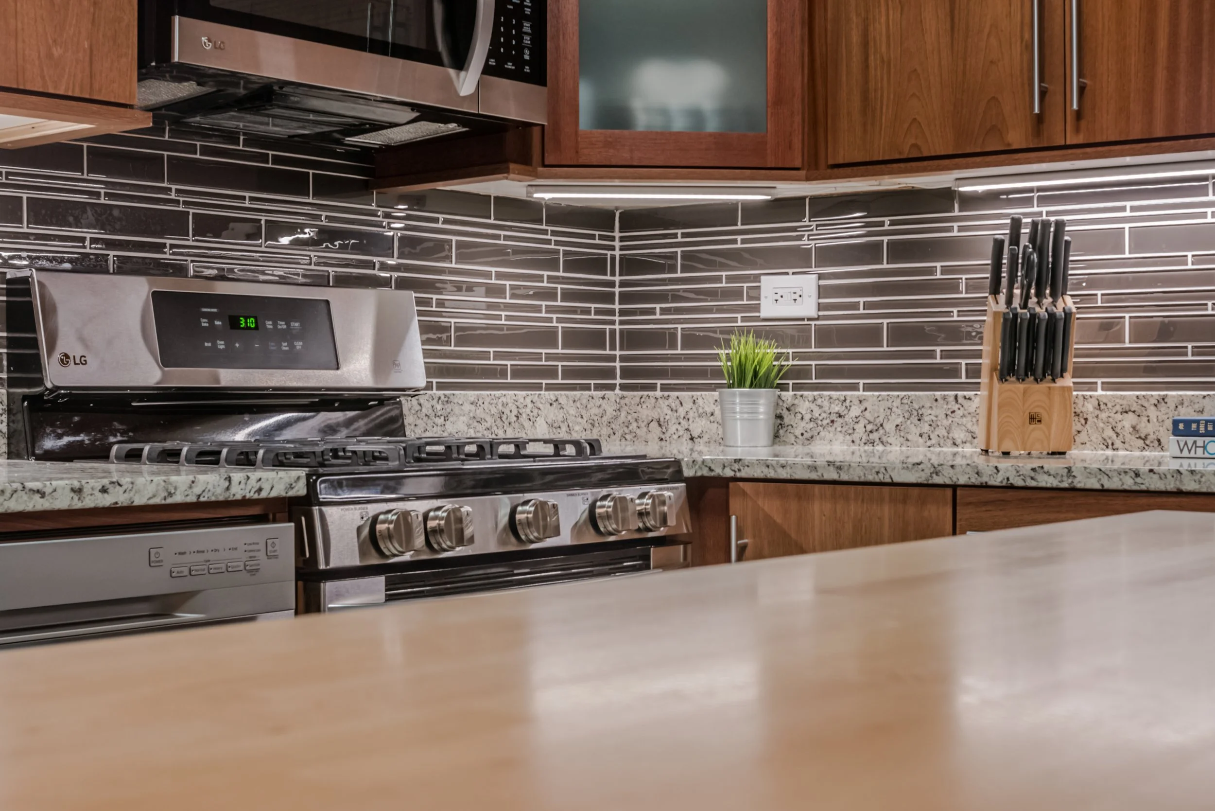 A modern kitchen with a stainless steel LG microwave over a stovetop, black and gray subway tile backsplash, wooden cabinets, a granite countertop, a small plant in a silver pot, a knife block, an electric outlet, and a box labeled "WHO".